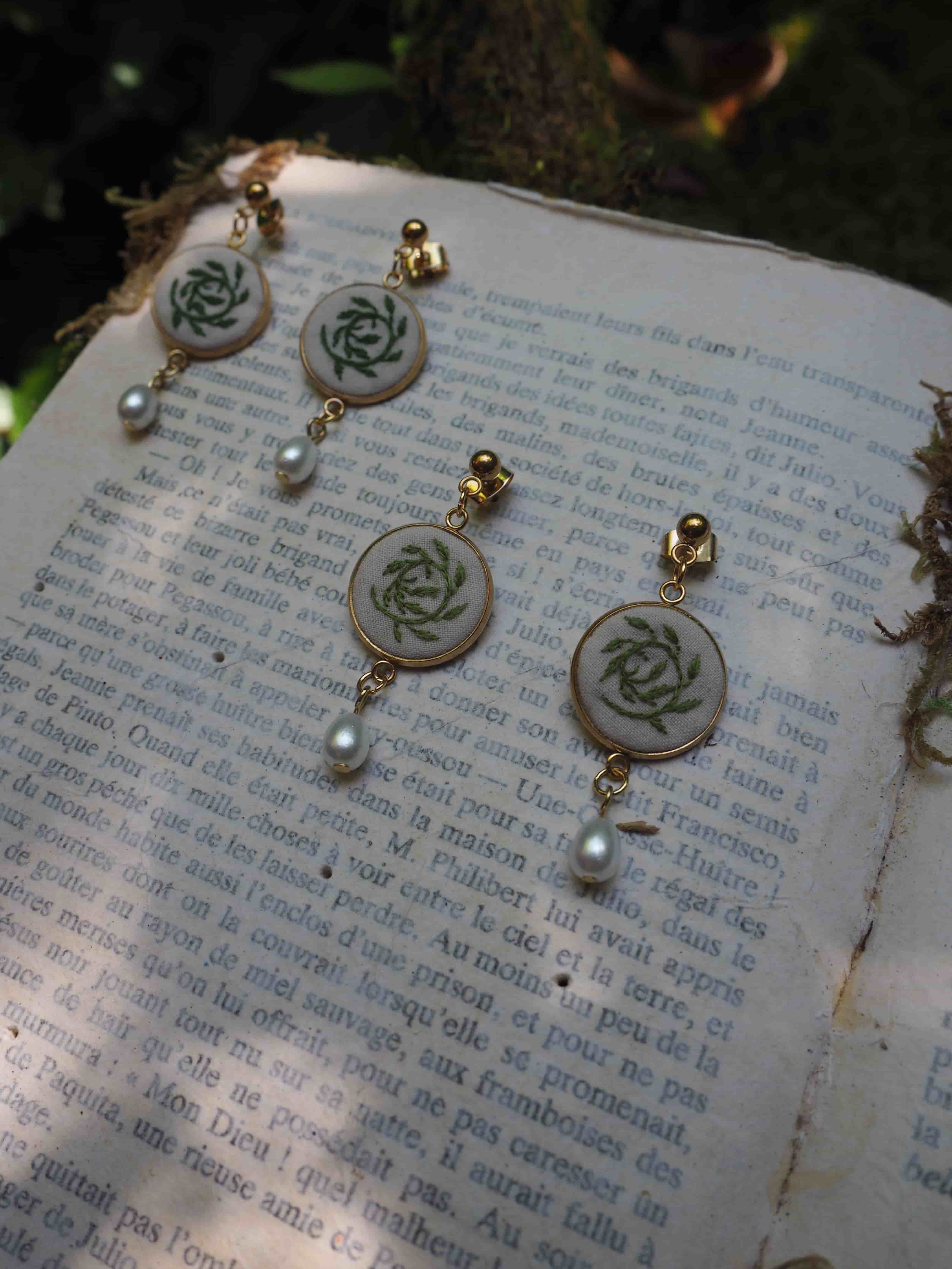 Cette image montre des bijoux délicats posés sur ce qui semble être une page de livre en français. Les bijoux sont des boucles d'oreilles qui présentent un motif de rose ou un motif floral circulaire en vert sur un fond clair, probablement de la porcelaine ou de l'émail. Chaque boucle d'oreille est ornée d'une petite perle blanche qui pend au bas. Les bijoux ont une apparence vintage ou artisanale, avec des détails dorés. Ils sont photographiés sur une page de texte qui semble être d'un livre ancien ou vintage, créant une composition romantique et nostalgique.