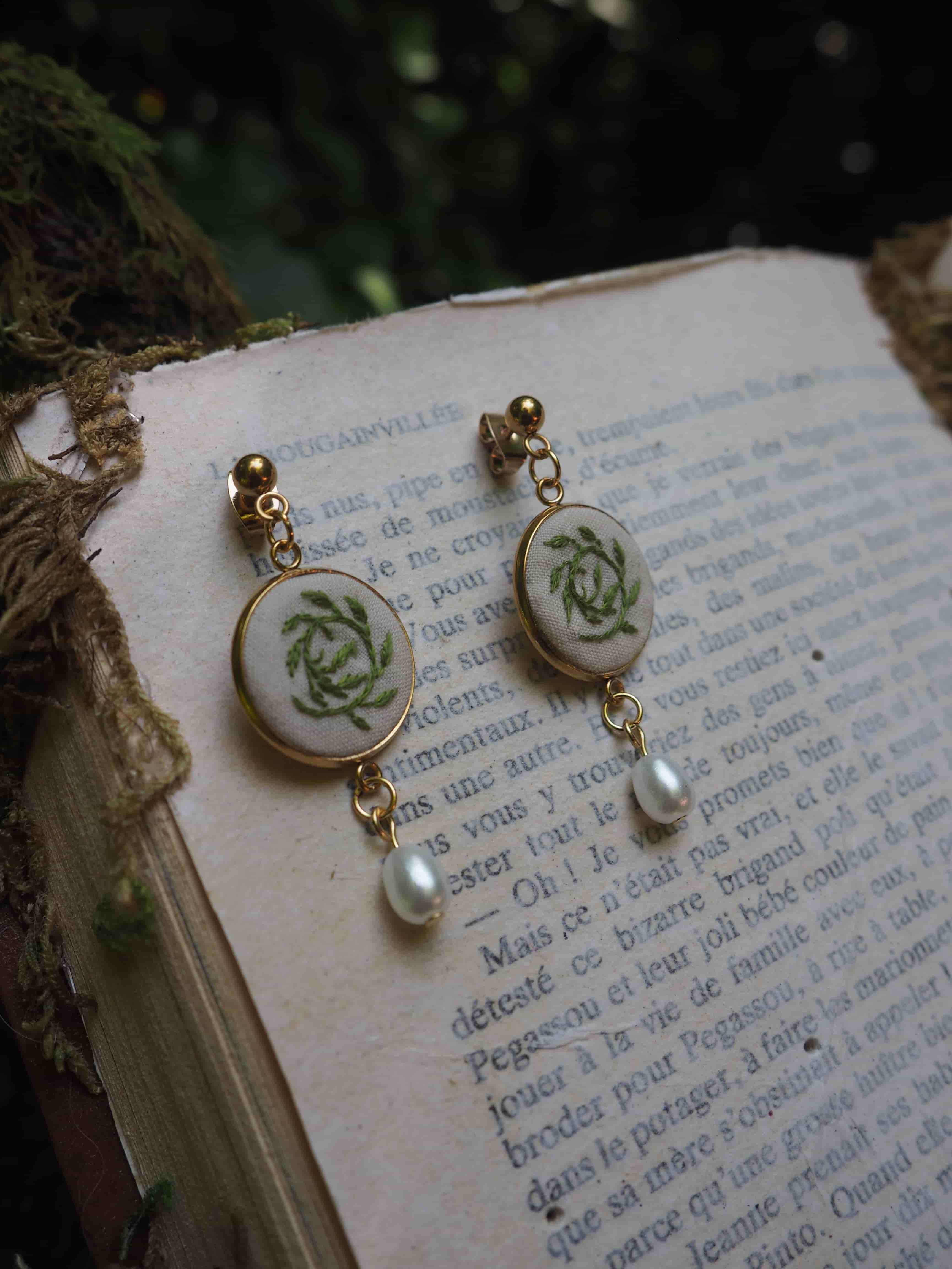 Cette image montre des boucles d'oreilles délicates posées sur ce qui semble être une page de livre en français. Les boucles d'oreilles paraissent être faites avec des petits médaillons en porcelaine ou en céramique, décorés de motifs floraux verts pâles, possiblement des roses. Elles sont agrémentées de perles et montées sur ce qui semble être des attaches dorées. Le fond montre du texte imprimé en français sur une page qui a un aspect vieilli ou vintage. L'image a une qualité romantique et vintage qui rappelle un style shabby chic ou français ancien.