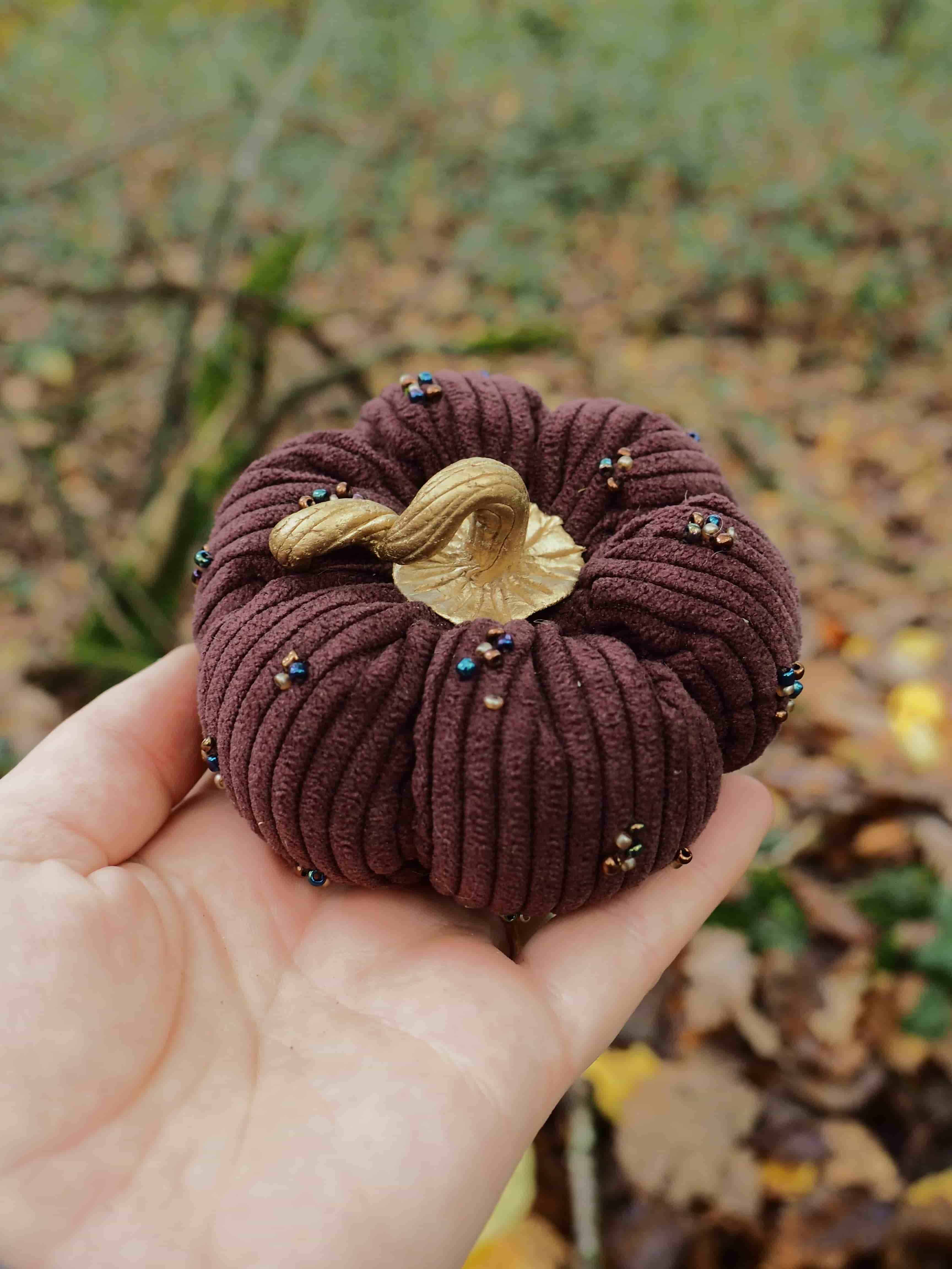 Cette image montre une main tenant délicatement un objet artisanal décoratif en forme de citrouille. L'objet présente les caractéristiques suivantes : **Forme et structure :**
- Forme ronde et cannelée typique d'une citrouille
- Texture côtelée créée par des plis réguliers
- Proportions compactes qui tiennent dans une paume de main **Matériaux et couleurs :**
- Tissu principal de couleur bordeaux/marron foncé
- Petites perles ou sequins dorés dispersés sur la surface
- Tige décorative dorée/beige au sommet **Contexte :**
- Photographié en extérieur sur un arrière-plan naturel flou
- Sol jonché de feuilles d'automne visibles
- Éclairage naturel qui met en valeur les détails de l'artisanat **Style :**
- Objet décoratif fait main, probablement pour la décoration d'automne
- Finition soignée avec attention aux détails
- Esthétique saisonnière évoquant Halloween ou Thanksgiving Il s'agit manifestement d'un élément de décoration artisanale de qualité, conçu pour célébrer la saison automnale.