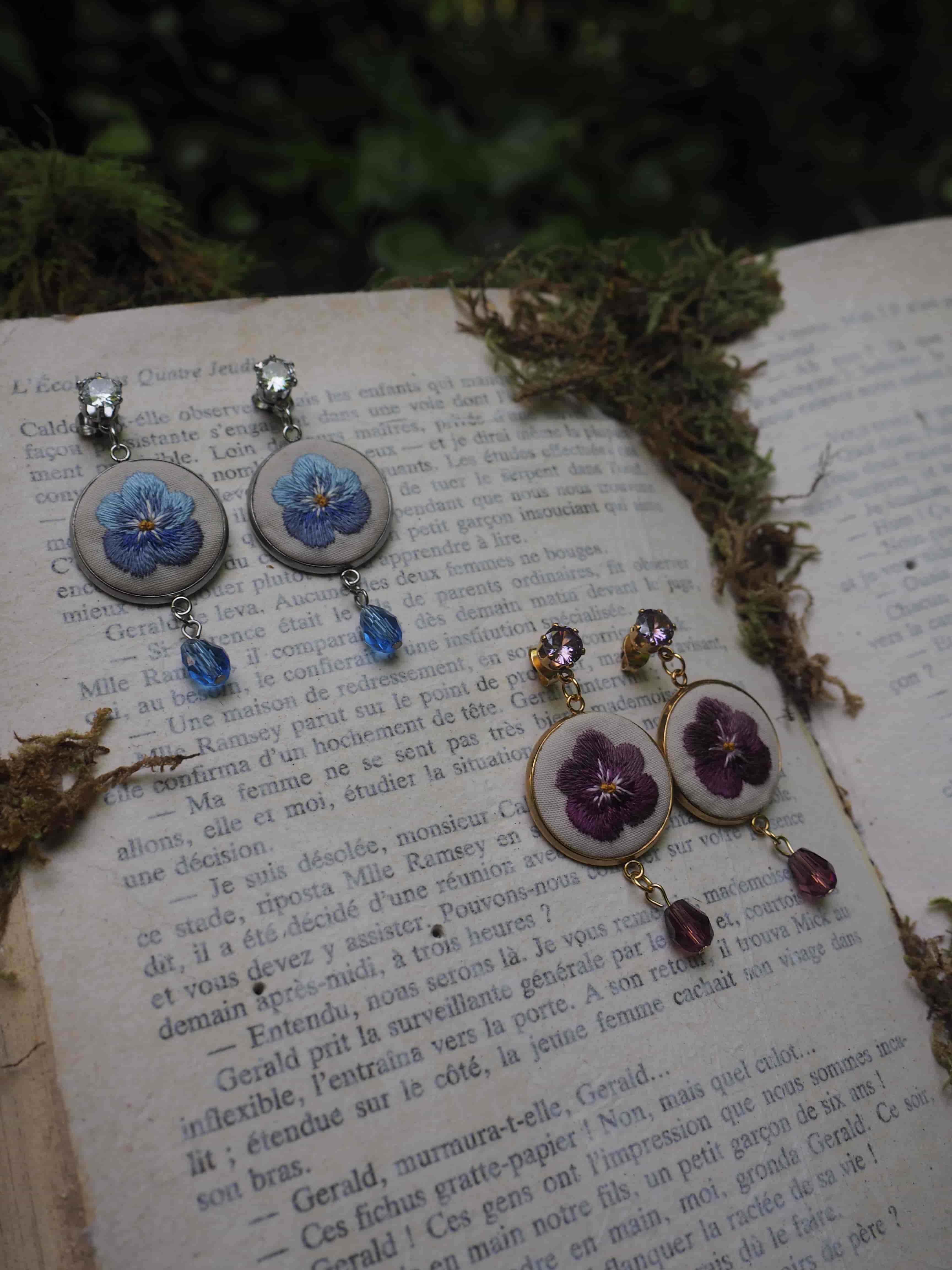 Cette image montre de jolies boucles d'oreilles artisanales posées sur les pages d'un livre ancien. Il y a deux paires de boucles d'oreilles : une paire avec des fleurs bleues et une autre avec des fleurs violettes. Les boucles d'oreilles semblent être faites dans un style vintage ou romantique, avec des pendentifs circulaires contenant les motifs floraux et des petites perles ou cristaux qui pendent en dessous. L'arrière-plan montre du texte imprimé sur les pages du livre et ce qui semble être de la mousse ou de la verdure sur le côté. La composition crée une ambiance très poétique et romantique, parfaite pour mettre en valeur ces bijoux délicats.