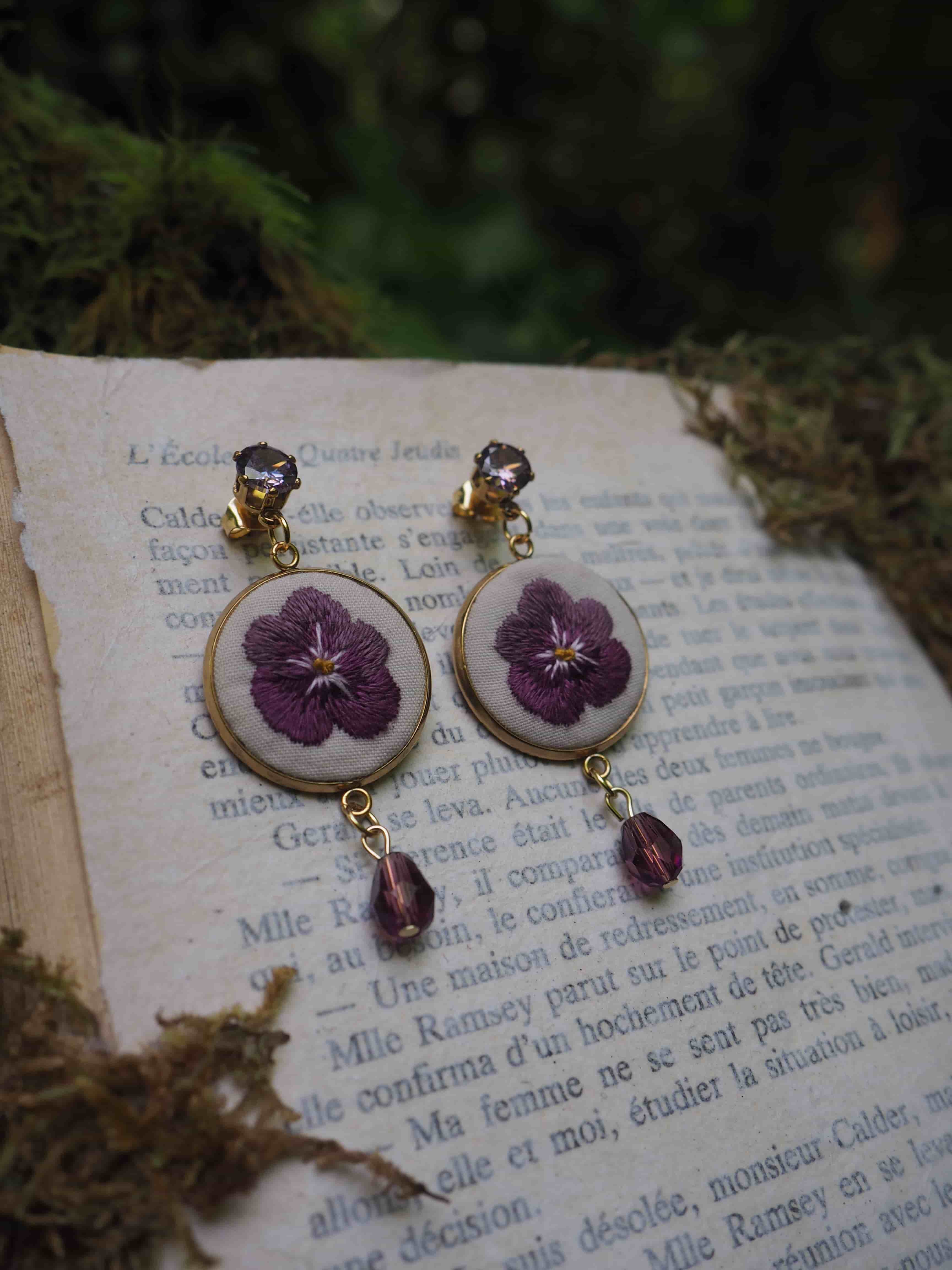 Ces boucles d'oreilles sont vraiment charmantes! Elles présentent de véritables fleurs pressées violettes (qui semblent être des pensées) préservées dans ce qui paraît être des supports circulaires blancs. Les boucles sont agrémentées de petites perles violettes qui pendent en dessous, créant un effet délicat et romantique. L'image est mise en scène sur ce qui semble être une page de livre en français, donnant une touche très poétique et vintage à la composition. Les tons violets des fleurs contrastent magnifiquement avec le fond blanc du support et la teinte jaunâtre de la page de livre. C'est un excellent exemple de bijoux artisanaux qui incorporent des éléments naturels de manière élégante.