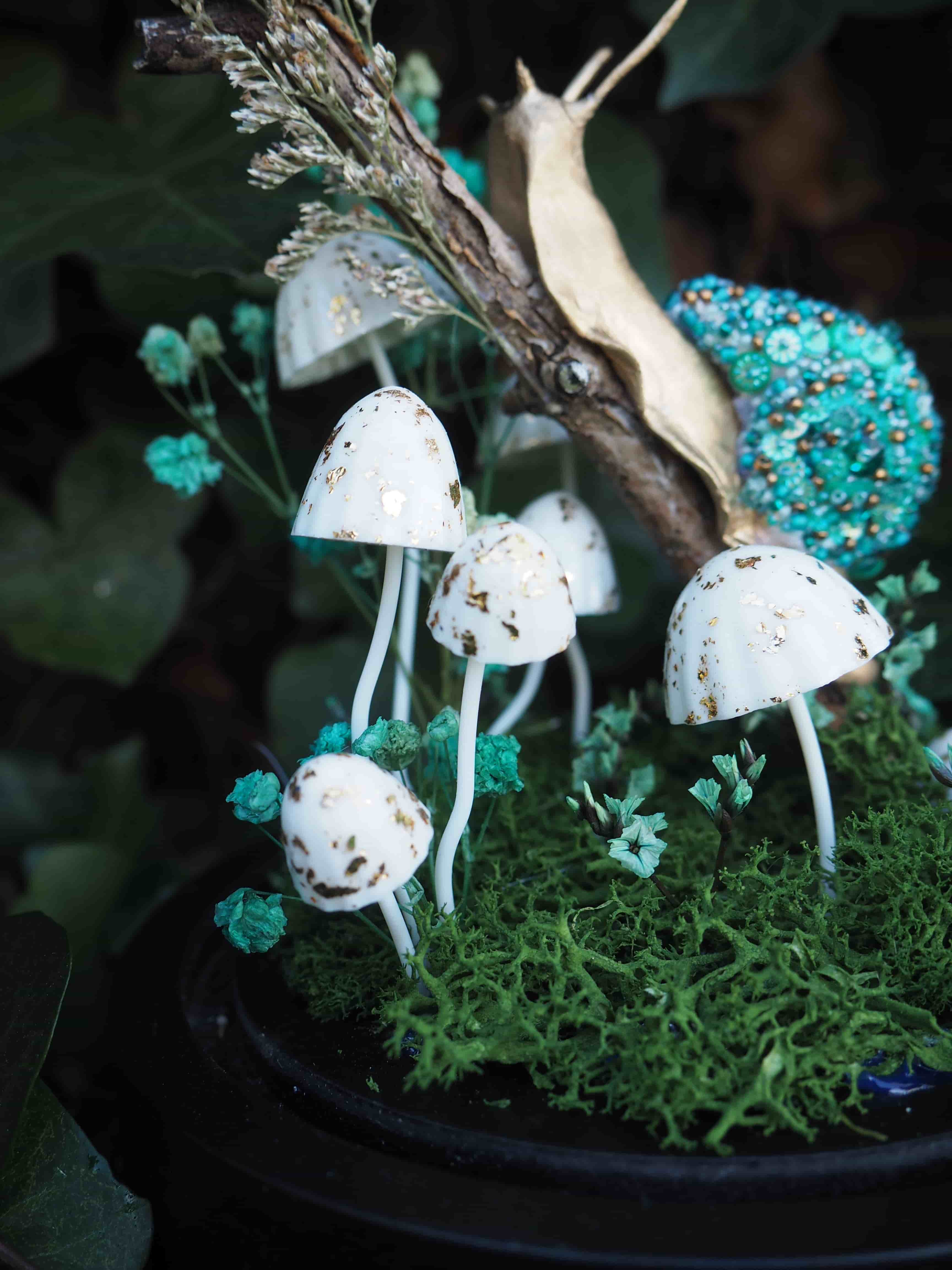 Cette image présente une scène miniature enchantée, centrée sur un arrangement de champignons et d'un escargot dans un environnement de mousse et de verdure. Au premier plan, on voit plusieurs petits champignons blancs avec des taches dorées, qui semblent être des décorations. Ils sont de tailles variées et sont plantés dans un lit de mousse verte texturée, qui occupe la majeure partie du bas de l'image. Entre les champignons et la mousse, on aperçoit de petites touffes de verdure d'un vert plus clair, presque turquoise, ajoutant de la profondeur et de la couleur à la scène. Au-dessus des champignons, un escargot stylisé est posé sur une branche ou un morceau de bois. La coquille de l'escargot est d'un bleu-vert vif, ornée de petites perles ou de paillettes qui lui donnent un aspect scintillant et texturé. Le corps de l'escargot est d'une couleur dorée ou beige clair, contrastant avec sa coquille. L'arrière-plan est flou, suggérant une profondeur de champ faible, ce qui met l'accent sur les éléments du premier plan. On distingue des teintes sombres de vert, probablement des feuilles ou de la végétation, qui contribuent à l'ambiance mystérieuse et forestière de la scène. L'ensemble de l'image dégage une atmosphère féerique et délicate, comme une petite scène de conte de fées ou un terrarium décoratif.