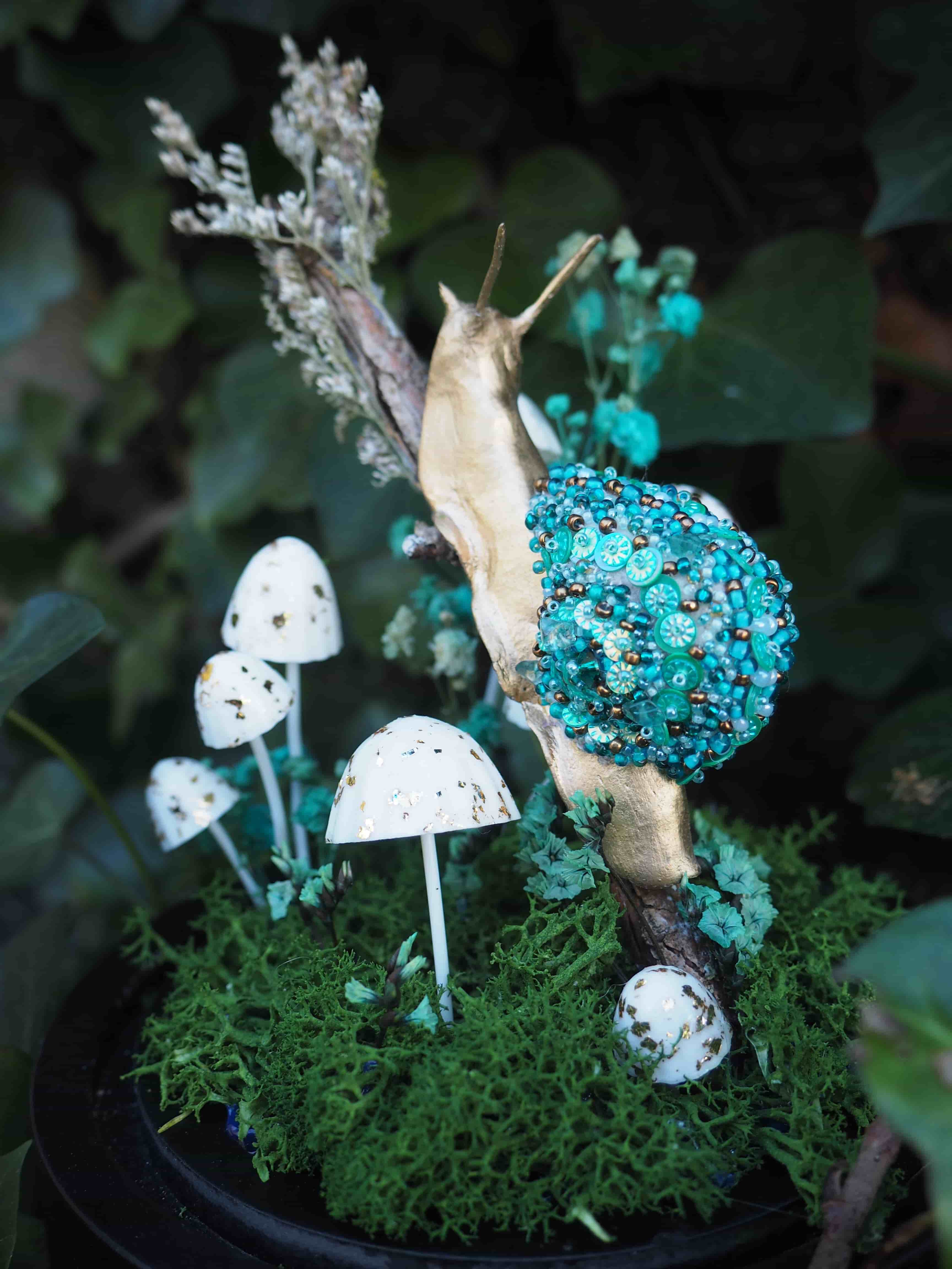 Cette image présente une composition artistique et fantaisiste, centrée sur une figurine d'escargot ornée, placée dans un décor miniature de forêt. Au premier plan, on voit un escargot stylisé, dont le corps est d'une couleur dorée ou laiton, lui donnant un aspect précieux. Sa coquille est particulièrement remarquable, recouverte d'une multitude de perles et de petites billes de couleur turquoise et bleu-vert, créant une texture riche et scintillante. L'escargot est positionné sur une branche ou un morceau de bois sec, qui semble être son support principal. Autour de l'escargot et à sa base, un tapis de mousse verte, dense et texturée, évoque un sous-bois. Plusieurs petits champignons miniatures, de couleur blanche avec des taches sombres (peut-être noires ou marron foncé), sont dispersés sur la mousse, ajoutant une touche de réalisme et de conte de fées à la scène. Certains de ces champignons ont des formes variées, certains étant plus élancés, d'autres plus trapus. En arrière-plan, on distingue un feuillage vert foncé et flou, suggérant un environnement naturel et luxuriant, comme un jardin ou une forêt. Quelques petites fleurs ou éléments végétaux de couleur bleu-vert pâle sont également visibles, ajoutant de la profondeur et de la couleur à l'ensemble. L'éclairage semble doux et naturel, mettant en valeur les détails et les textures des différents éléments. L'ensemble de la composition est probablement une œuvre d'art décorative ou un diorama, conçu pour évoquer un monde miniature enchanté.