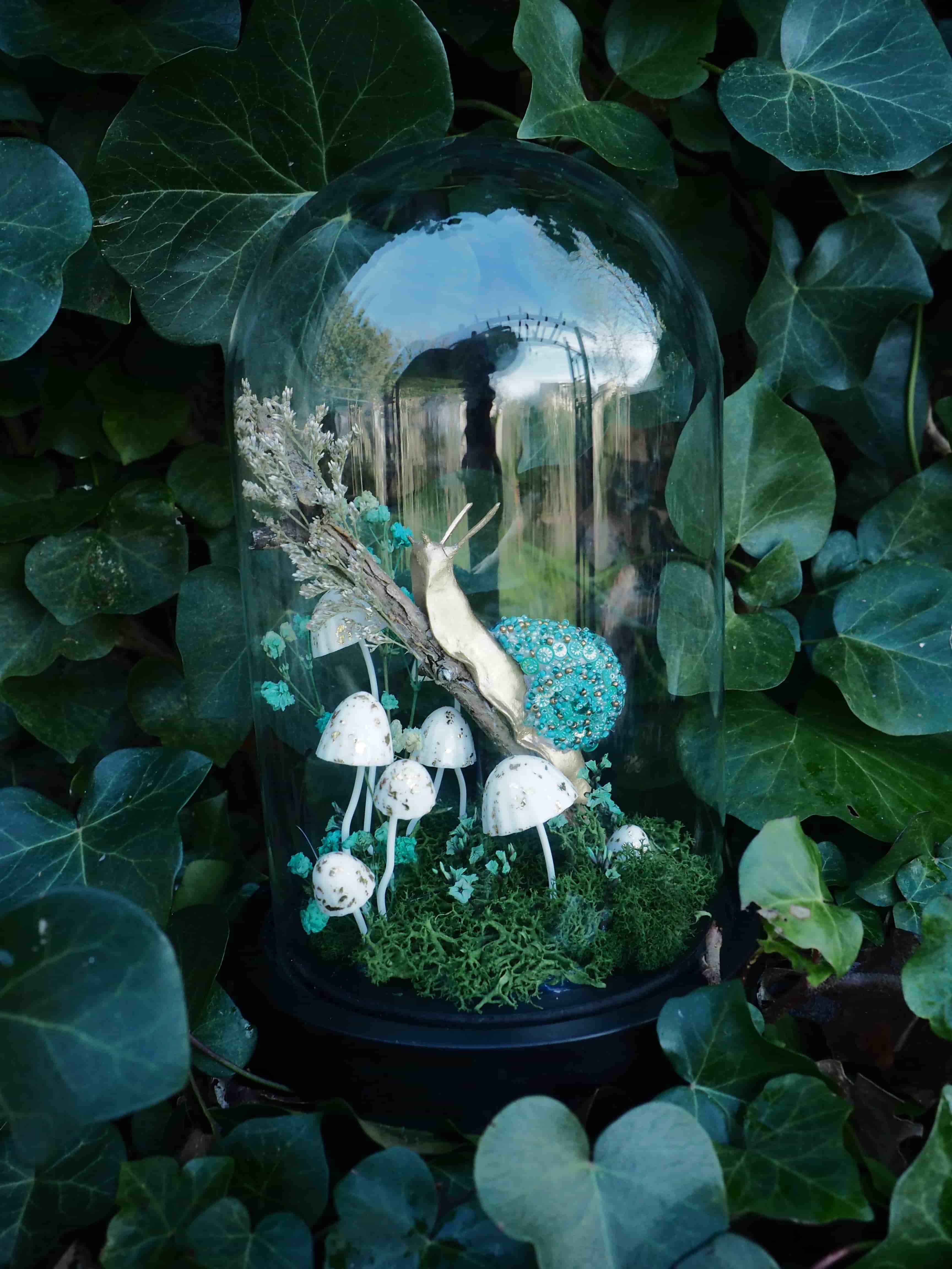 L'image présente un terrarium en forme de cloche en verre, posé au milieu d'un feuillage vert luxuriant, probablement du lierre. À l'intérieur de la cloche, on observe une scène miniature de forêt ou de sous-bois. Au centre, un escargot de couleur claire, presque blanc ou beige, est posé sur une branche ou une brindille. Autour de lui, plusieurs champignons blancs avec des chapeaux ronds et des pieds fins sont disposés, créant une ambiance féerique. Le sol du terrarium est recouvert de mousse verte, et on peut distinguer quelques éléments décoratifs bleus ou turquoise, peut-être des petites pierres ou des fragments de verre coloré, ajoutant une touche de couleur. À l'arrière-plan, il y a aussi des éléments végétaux secs ou des herbes hautes de couleur claire. La cloche en verre reflète légèrement l'environnement extérieur, et la lumière semble venir d'en haut, illuminant la scène intérieure. L'ensemble dégage une atmosphère mystérieuse et naturelle, comme un petit écosystème préservé.