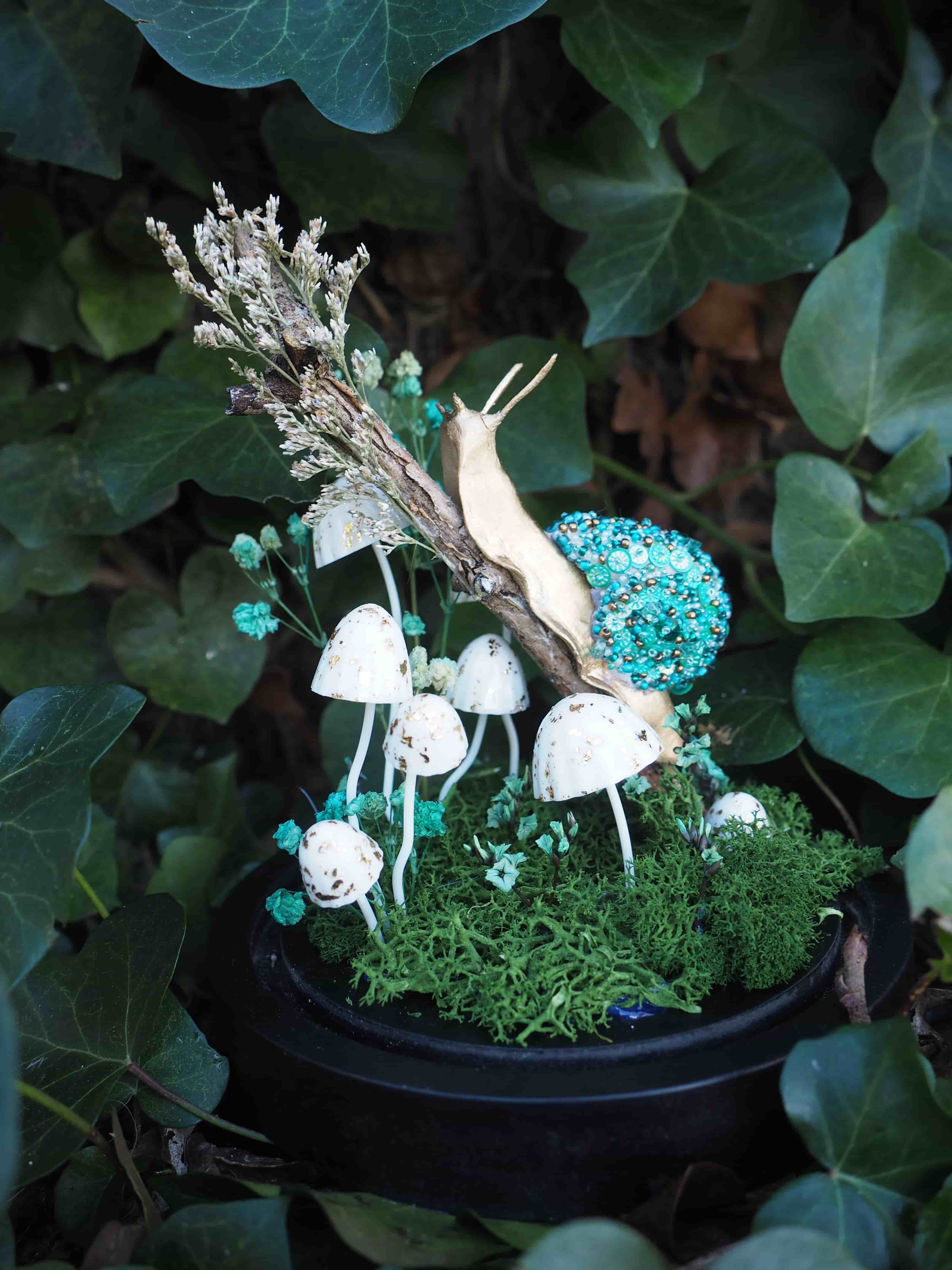 L'image présente une composition artistique et fantaisiste, évoquant un petit écosystème miniature ou un terrarium, avec un thème forestier et féerique. Au centre de l'image, un escargot doré, stylisé et brillant, est positionné sur une branche ou un morceau de bois sec. Sa coquille est ornée de petites perles ou de paillettes bleues-vertes scintillantes, lui donnant un aspect précieux et irréel. Sous l'escargot et la branche, un groupe de petits champignons blancs, aux chapeaux ronds et aux pieds fins, pousse sur un lit de mousse verte luxuriante. Certains champignons sont plus grands et bien visibles, tandis que d'autres sont plus petits et se fondent davantage dans la mousse. La mousse verte forme un tapis dense et texturé, créant un sol naturel pour cette scène miniature. Quelques petites fleurs ou éléments décoratifs de couleur bleu-vert clair sont parsemés dans la mousse et autour des champignons, ajoutant une touche de couleur et de délicatesse. L'ensemble de cette composition est contenu dans un récipient sombre, probablement un pot ou une coupelle noire, qui sert de base et de contraste aux couleurs vives de la scène. L'arrière-plan est flou et dominé par des feuilles de lierre vert foncé, créant une ambiance naturelle et mystérieuse, comme si cette scène miniature était cachée au cœur d'une forêt. La lumière semble douce et diffuse, mettant en valeur les détails et les textures de chaque élément. L'image dégage une atmosphère de conte de fées, de magie et de nature enchantée, avec une attention particulière aux détails et à l'esthétique.