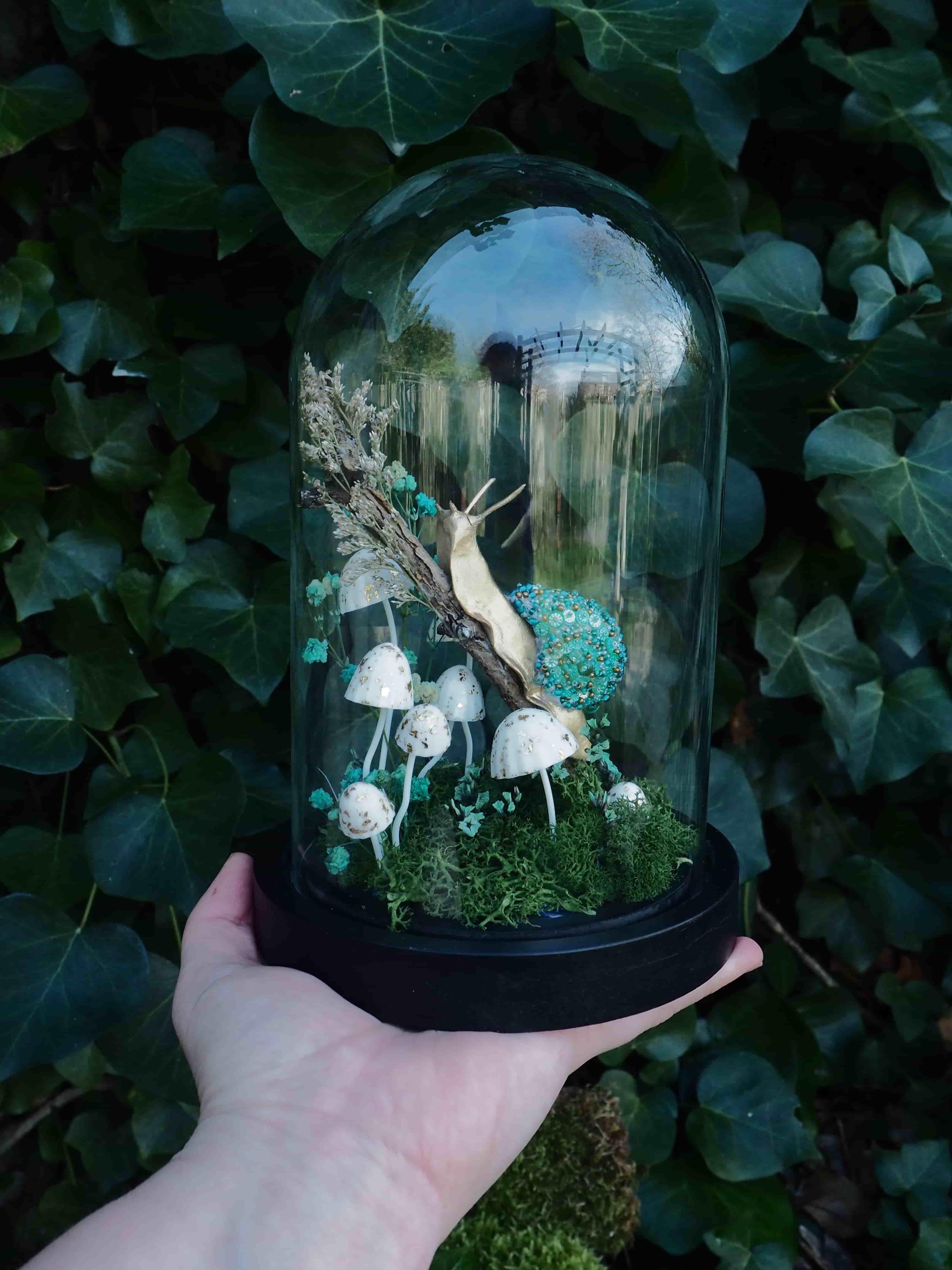 This image features a close-up shot of a person's hand holding a glass cloche, which encases a miniature, whimsical diorama. The cloche has a black base and a clear, domed glass cover. Inside the cloche, a fantastical scene unfolds:
*   **Central Figure:** A prominent, elongated snail-like creature with a light, almost golden-brown body and a vibrant, textured, teal-colored shell is positioned towards the upper center. It appears to be climbing or resting on a piece of dried, light-colored foliage or wood.
*   **Mushrooms:** Several small, white-capped mushrooms with slender stems are scattered across the base of the diorama. They vary slightly in size and arrangement, adding to the enchanted forest aesthetic.
*   **Foliage/Moss:** The base of the diorama is covered with lush, green moss, creating a natural ground cover. There are also some dried, lighter-colored plant elements, possibly twigs or dried flowers, contributing to the texture and depth of the scene.
*   **Background Elements:** In the background within the cloche, through the glass, there are hints of what appears to be a miniature bridge or structure, suggesting a larger, imagined landscape. The hand holding the cloche is light-skinned, with visible fingers and palm, providing a sense of scale to the miniature artwork. The background outside the cloche is dominated by dark green ivy leaves, which are slightly out of focus, emphasizing the cloche and its contents. The lighting appears natural, highlighting the details within the glass dome. Overall, the image conveys a sense of delicate craftsmanship and a magical, miniature world contained within the glass.