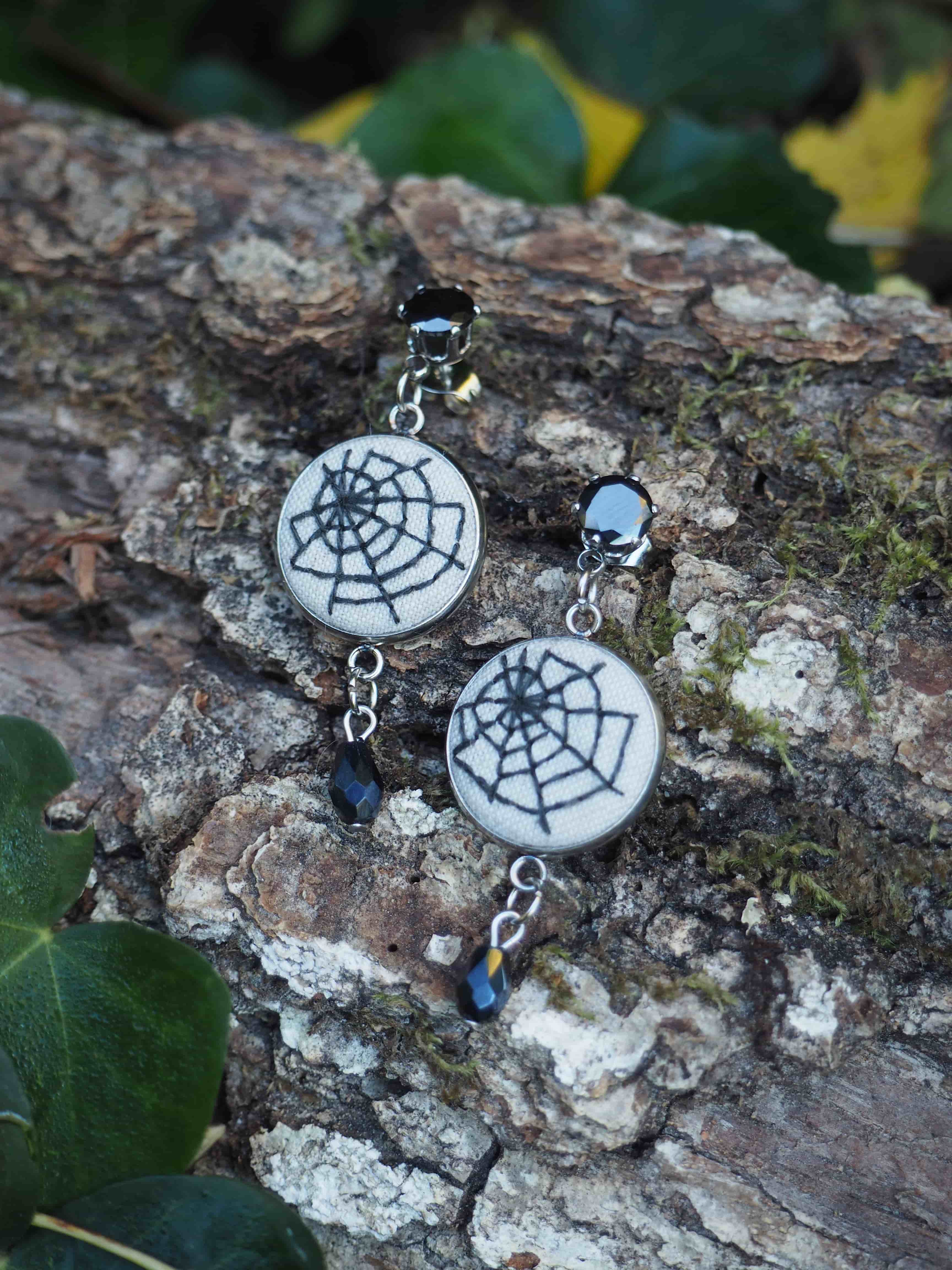 Cette image présente une paire de boucles d'oreilles artisanales disposées sur une surface naturelle en écorce d'arbre. Les bijoux arborent un design de toile d'araignée en métal argenté, créant un motif géométrique délicat et symétrique. Chaque boucle d'oreille est ornée de petites perles bleues qui ajoutent une touche de couleur subtile à l'ensemble. Les boucles d'oreilles sont photographiées dans un environnement naturel, posées sur l'écorce rugueuse et texturée d'un tronc d'arbre. On peut apercevoir en arrière-plan un feuillage vert flou qui crée une ambiance organique et naturelle, mettant en valeur le contraste entre l'artisanat métallique et l'environnement forestier. L'éclairage naturel met en relief les détails du travail métallique et fait ressortir les nuances de couleur des perles bleues. Cette mise en scène suggère un style bohème ou naturel, évoquant peut-être des bijoux inspirés de la nature ou de thèmes mystiques liés aux toiles d'araignée.