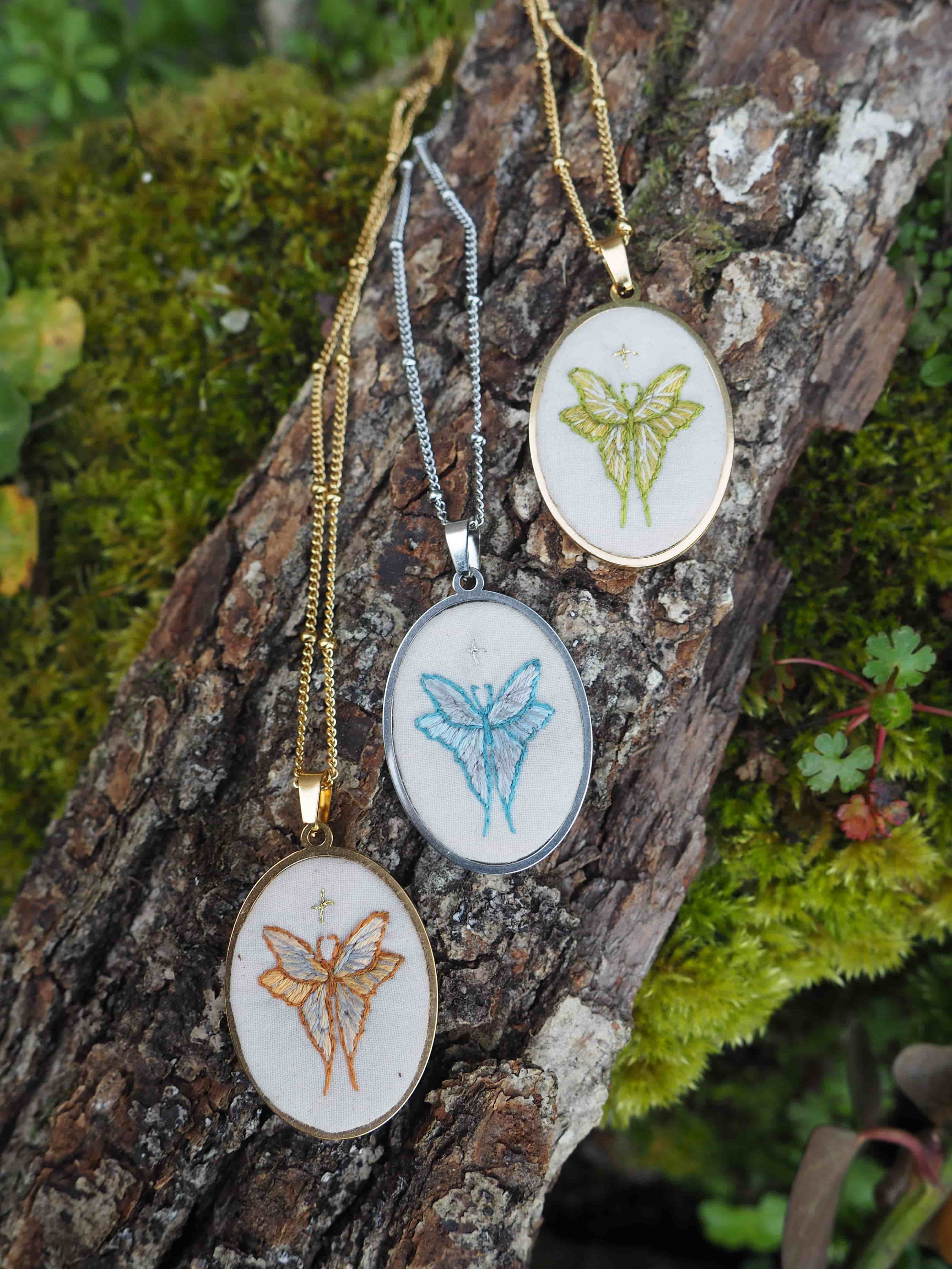 Cette image montre trois jolis pendentifs brodés disposés sur un morceau d'écorce couvert de mousse. Chaque pendentif présente un papillon brodé dans des couleurs différentes - l'un en jaune/vert, un autre en bleu clair, et le troisième en orange/doré. Les pendentifs sont montés sur des chaînes de différentes couleurs assorties (dorée et argentée) et semblent être des pièces de bijouterie artisanale délicate. Le cadre naturel avec l'écorce moussue et la verdure en arrière-plan met parfaitement en valeur ces créations délicates. C'est une belle composition qui marie l'artisanat et la nature.