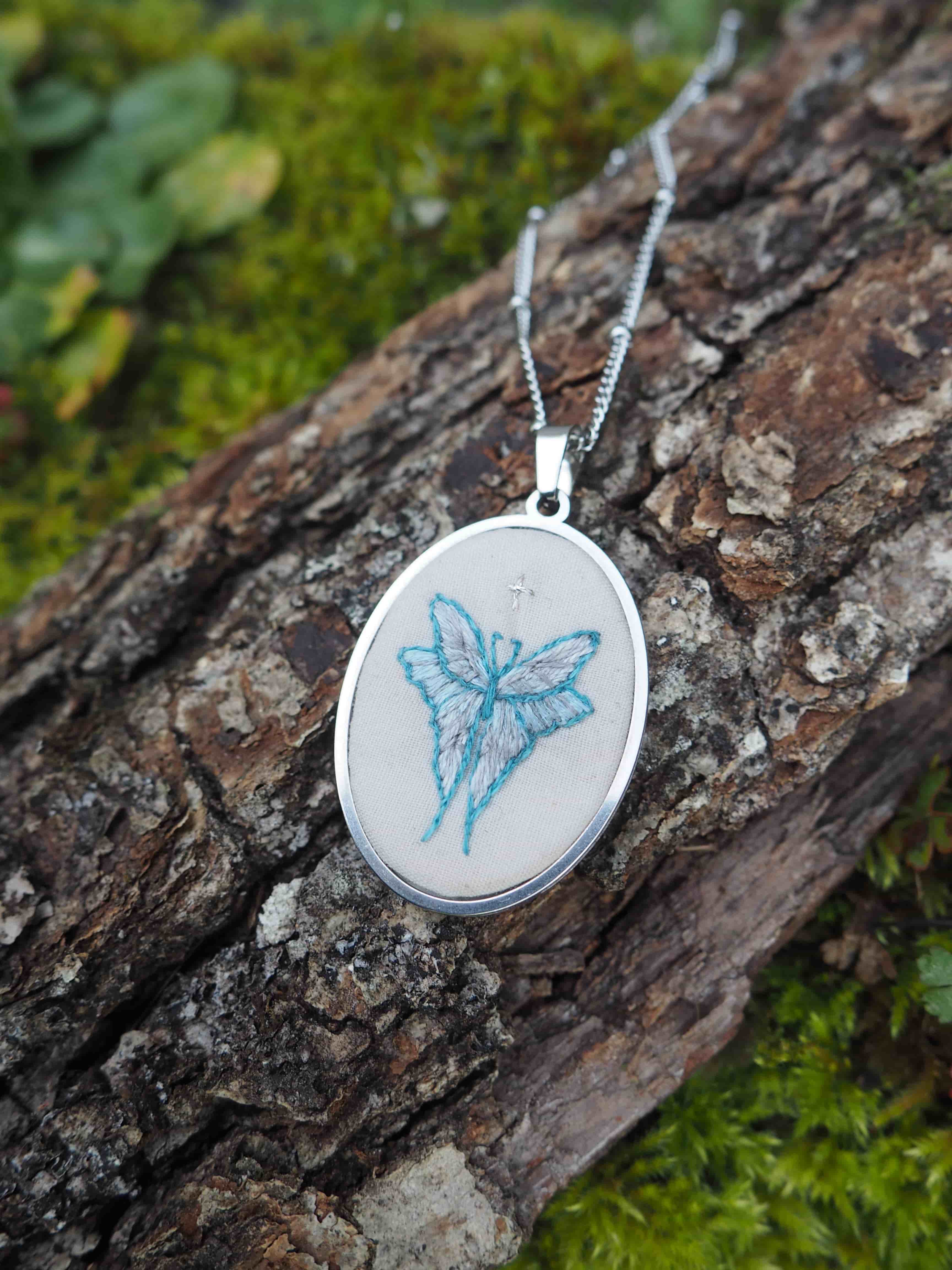 Cette image montre un pendentif délicat sur une chaîne en argent, photographié sur un morceau d'écorce d'arbre couvert de mousse verte. Le pendentif est rond et contient une broderie ou un dessin d'un papillon bleu sur un fond blanc. Le design est simple mais élégant, avec le papillon représenté dans des tons bleu clair. Le contraste entre le bijou argenté et la texture naturelle rugueuse de l'écorce, ainsi que la mousse verte, crée une belle composition photographique qui met en valeur la délicatesse du pendentif.
