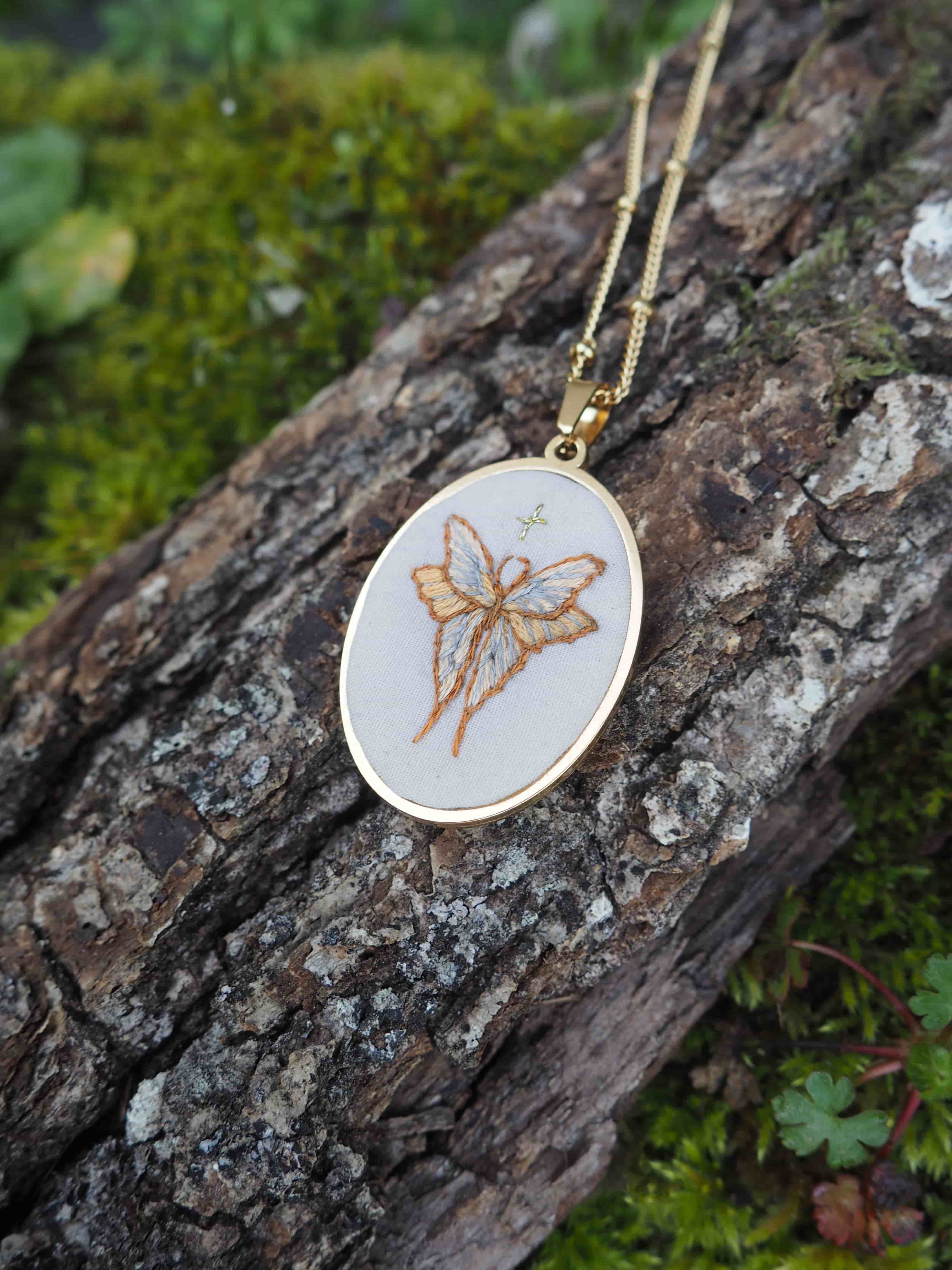 Cette image montre un délicat pendentif artisanal composé d'un cercle à broder (tambour de broderie miniature) monté en collier avec une chaîne dorée. Le motif brodé représente un papillon dans des tons dorés ou beiges sur un fond blanc. Le pendentif est photographié sur un tronc d'arbre recouvert de mousse verte, ce qui crée un beau contraste naturel avec le bijou. C'est une pièce de bijouterie très délicate et naturelle qui allie l'art de la broderie à la joaillerie.