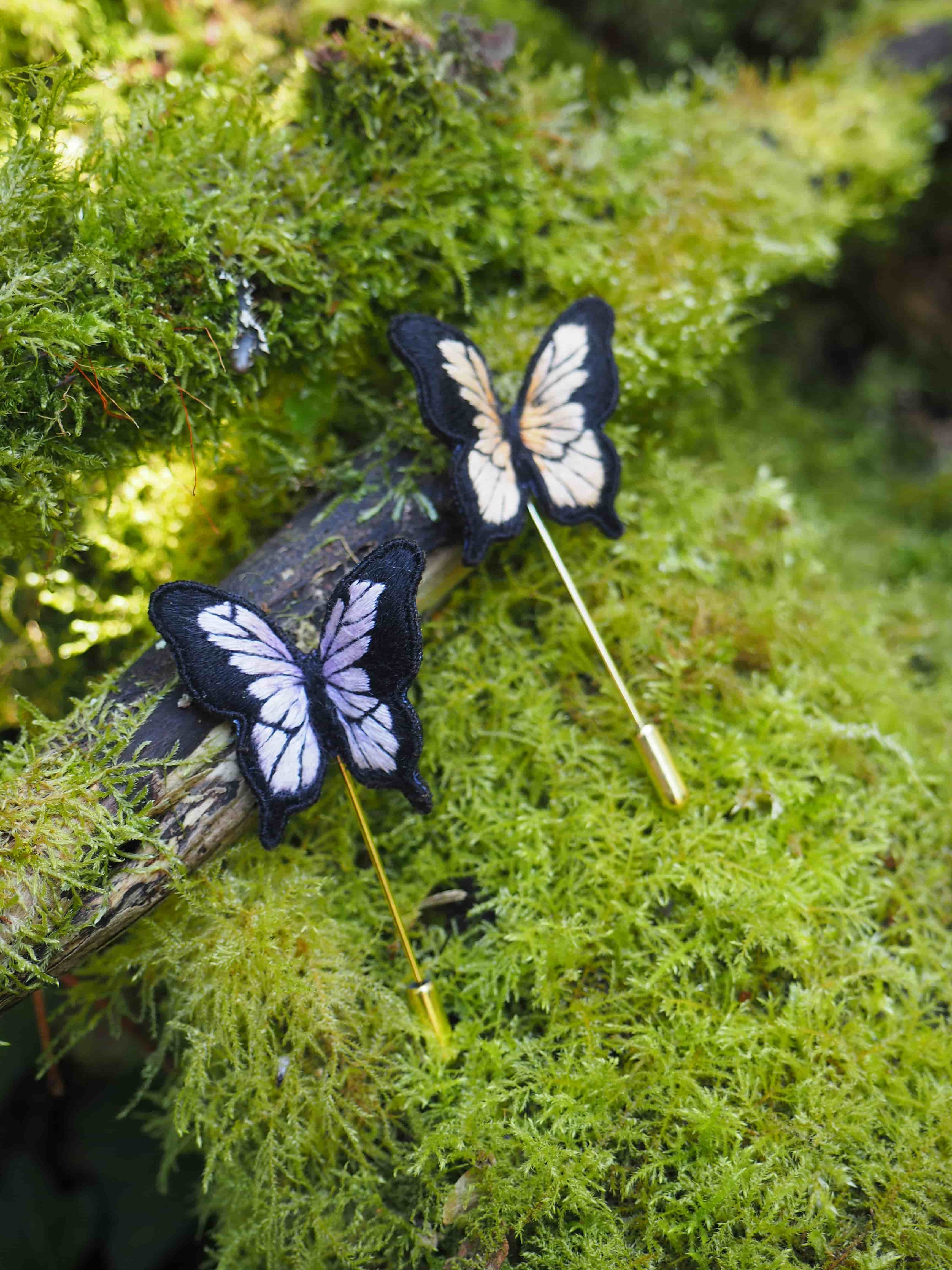 Cette image montre deux jolies décorations en forme de papillons posées sur un lit de mousse verte luxuriante. Les papillons sont de couleur bleu foncé et blanc, probablement faits à la main, et semblent être montés sur de fines tiges. Le contraste entre les papillons bleu-blanc et le vert vif de la mousse crée une scène très naturelle et apaisante. La mousse qui sert d'arrière-plan a une texture douce et vaporeuse, donnant à l'ensemble une ambiance de jardin féerique ou de sous-bois enchanteur.