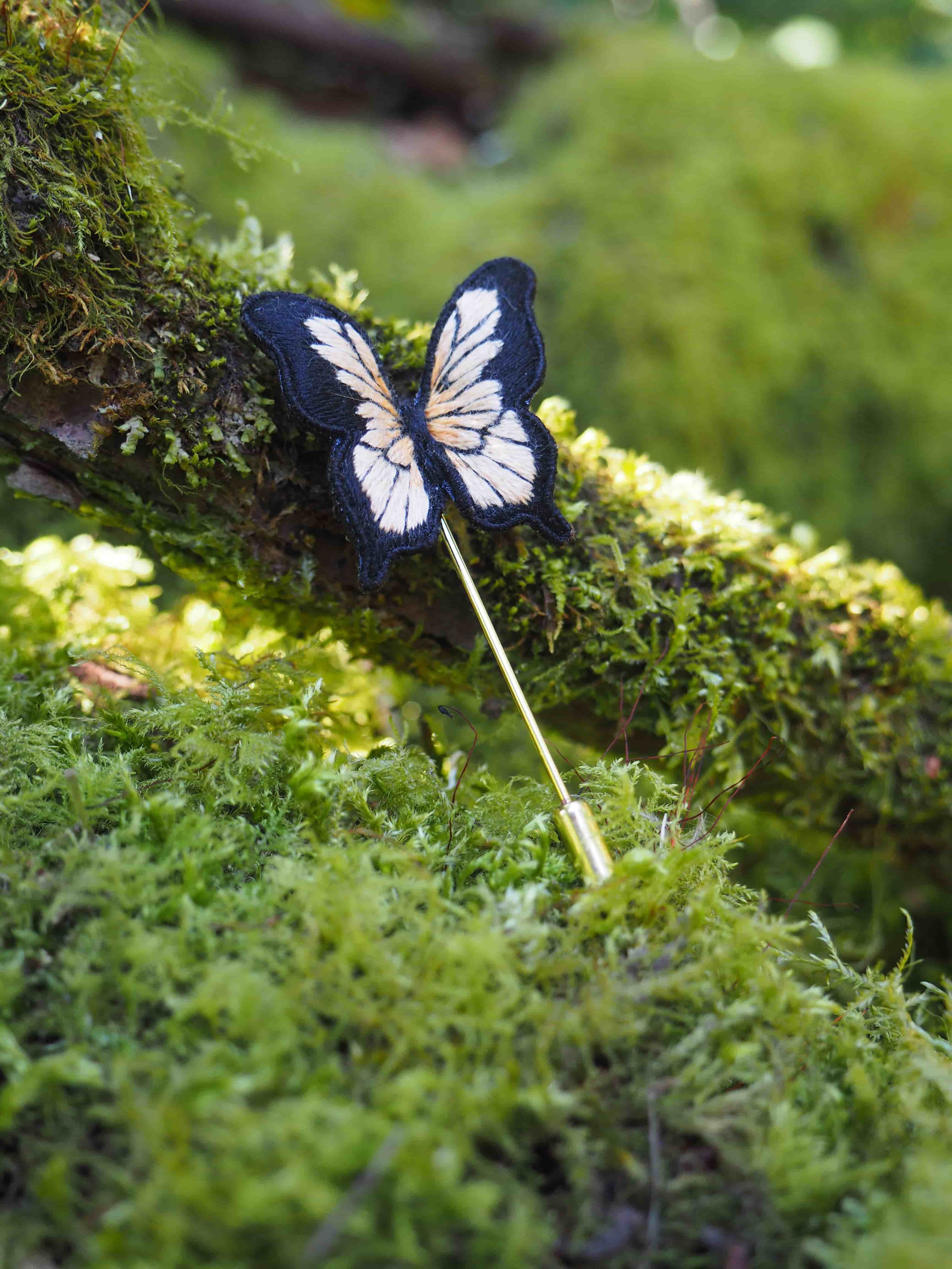 Cette image montre une délicate broche ou épingle décorative en forme de papillon, posée sur un lit de mousse verte. Le papillon est de couleur bleu foncé et blanc, avec des ailes finement détaillées qui rappellent celles d'un papillon naturel. L'épingle est fixée sur une longue tige fine, et l'ensemble crée un beau contraste avec le fond vert et texturé de la mousse. La photo a une ambiance très naturelle et poétique, avec une belle profondeur de champ qui met en valeur le papillon tout en gardant un arrière-plan légèrement flou. La lumière douce qui éclaire la scène donne un aspect féerique à l'ensemble.