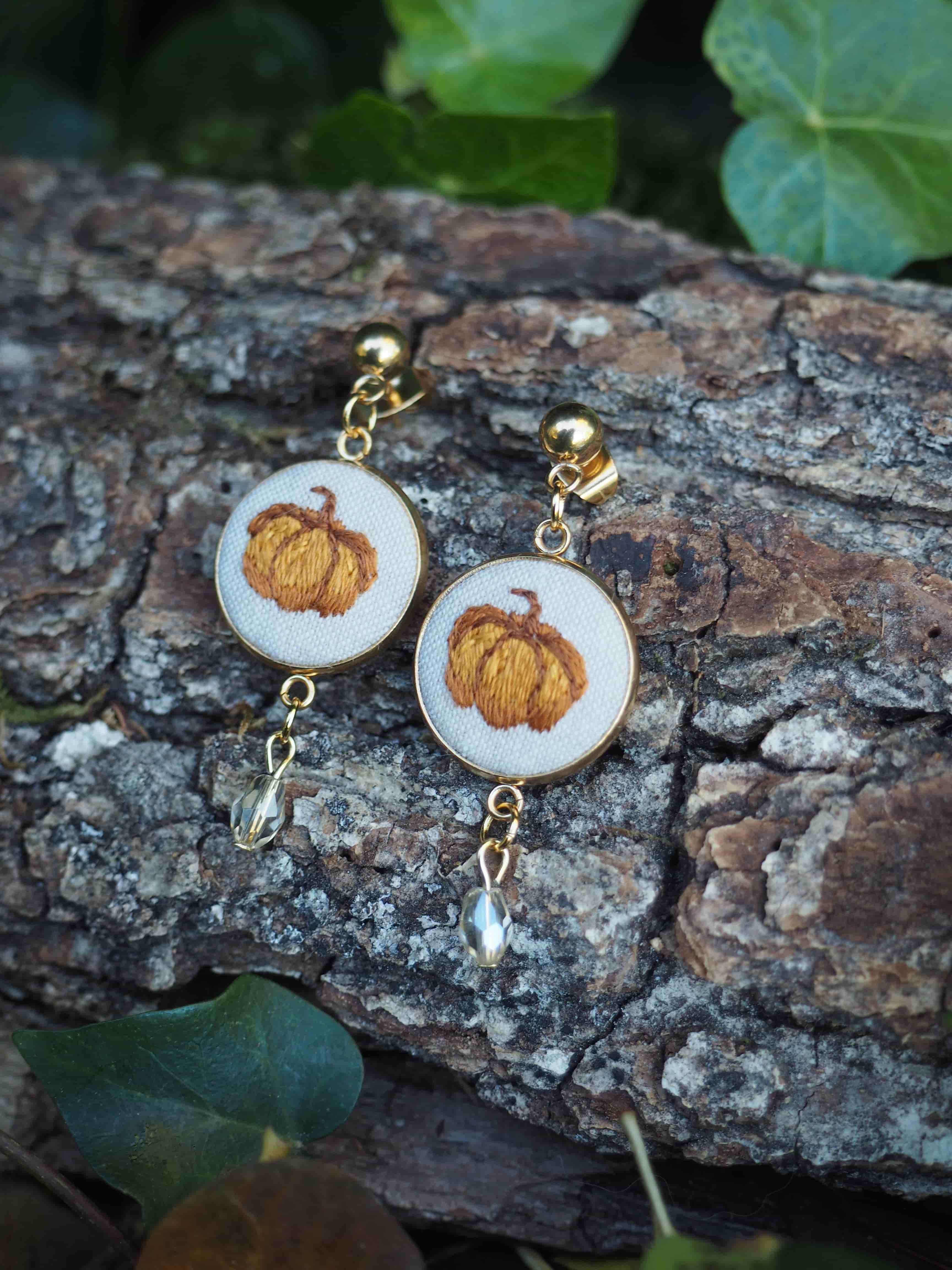 Cette image présente une paire de boucles d'oreilles artisanales disposées sur un morceau d'écorce naturelle dans un environnement végétal. **Caractéristiques des bijoux :**
- Pendants circulaires en résine ou matériau translucide blanc/nacré
- Inclusion de véritables feuilles d'automne aux teintes dorées/orangées au centre de chaque cercle
- Montures et crochets en métal doré
- Petites perles dorées comme éléments décoratifs suspendus **Mise en scène :**
- Support naturel : morceau d'écorce rugueuse aux tons bruns et gris
- Environnement : feuillage vert en arrière-plan, créant un contraste naturel
- Éclairage doux qui fait ressortir les reflets dorés des bijoux **Style :**
Ces boucles d'oreilles s'inscrivent dans une esthétique bohème et naturelle, mêlant éléments organiques (vraies feuilles) et matériaux modernes (résine). Elles évoquent l'automne et la beauté de la nature préservée dans un bijou délicat. La photographie met en valeur le caractère artisanal et poétique de ces créations bijoutières.