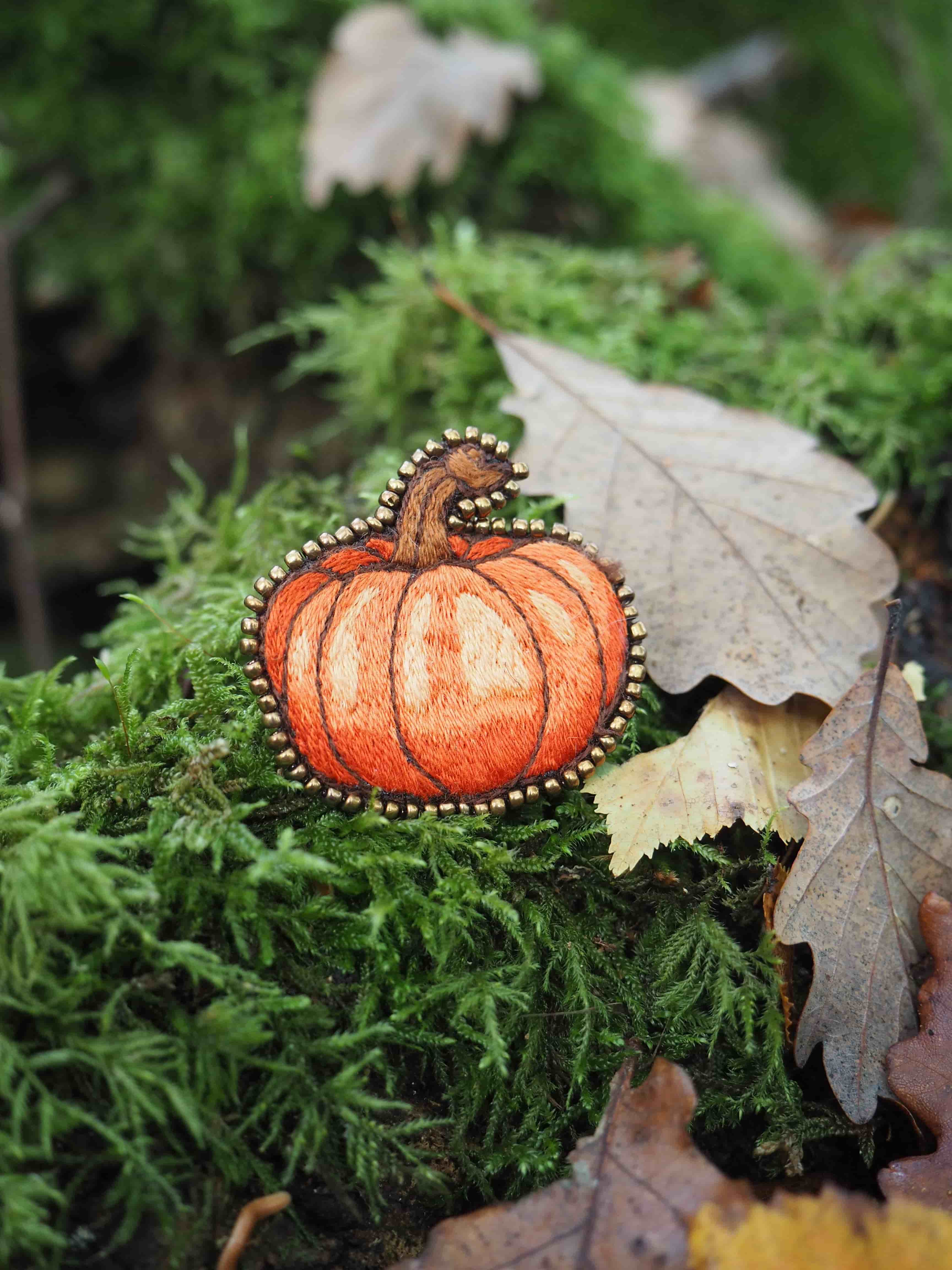 Cette image présente une scène automnale charmante et artistique. Au centre, on peut voir un petit pendentif ou une broche décorative en forme de citrouille orange, finement détaillée avec des contours perlés ou brodés qui lui donnent un aspect artisanal élégant. La citrouille miniature repose délicatement sur un tapis de mousse verte luxuriante, créant un beau contraste de couleurs entre l'orange vif du motif et le vert naturel de la mousse. À côté, on aperçoit des feuilles mortes aux tons bruns et beiges, typiques de la saison automnale. L'ensemble évoque une ambiance forestière paisible, avec la mousse qui suggère un environnement humide et ombragé. La composition photographique met l'accent sur les textures naturelles - la douceur de la mousse, la rugosité des feuilles séchées - tout en mettant en valeur ce petit objet décoratif qui semble célébrer l'esprit d'Halloween ou de l'automne. L'éclairage doux et la profondeur de champ créent une atmosphère intimiste et poétique, transformant cette simple nature morte en une véritable œuvre artistique.