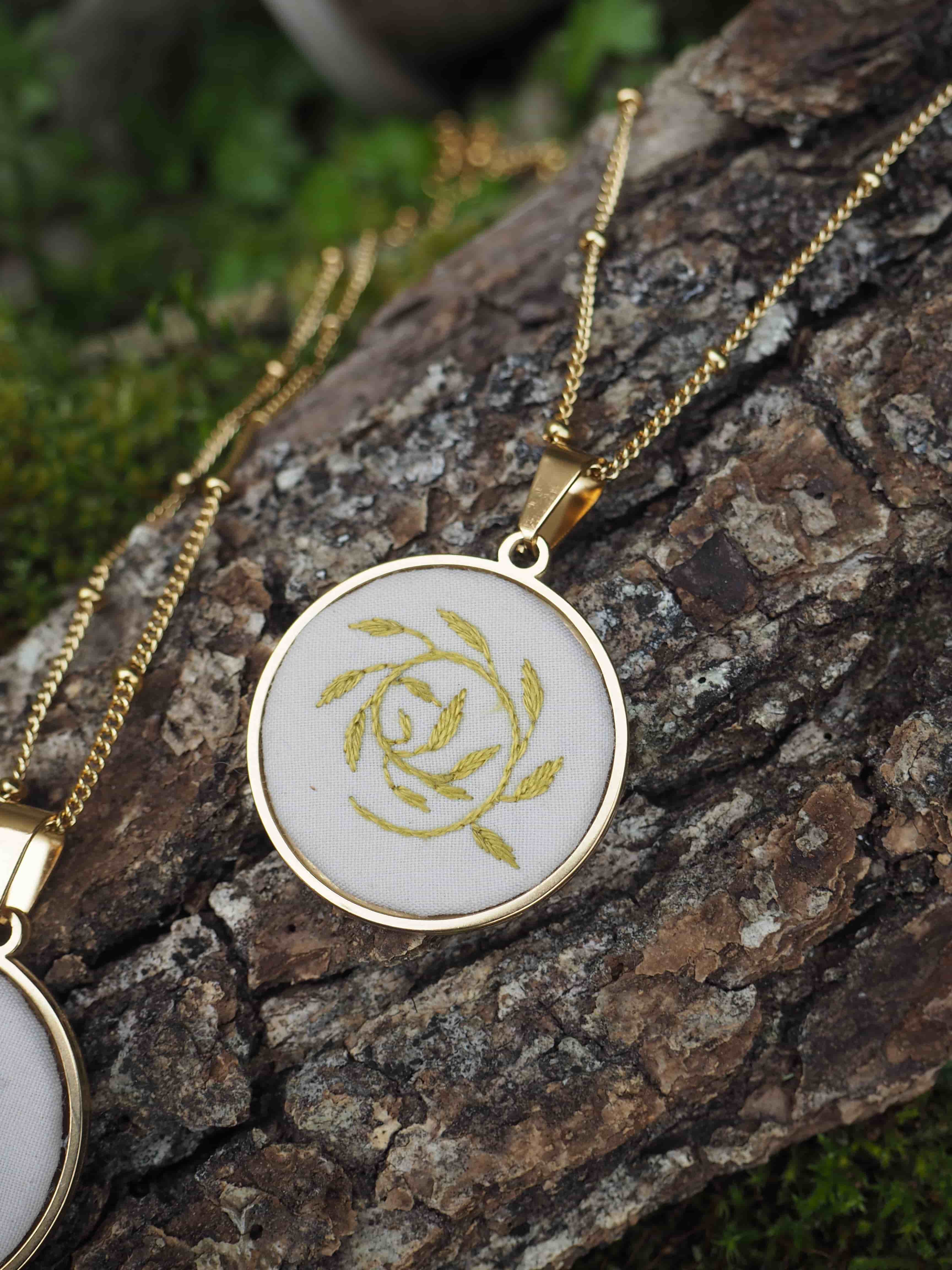 Cette image montre un pendentif délicat sur une chaîne dorée, posé sur un morceau de bois couvert de mousse. Le pendentif est de forme ovale avec un cadre doré et contient ce qui semble être une broderie ou un motif de rose stylisée en fil doré sur un fond blanc ou crème. La photographie est prise en gros plan avec une belle profondeur de champ, mettant en valeur les détails du bijou tout en montrant la texture naturelle du bois et la mousse verte en arrière-plan. C'est une composition élégante qui marie les éléments naturels avec un bijou raffiné.