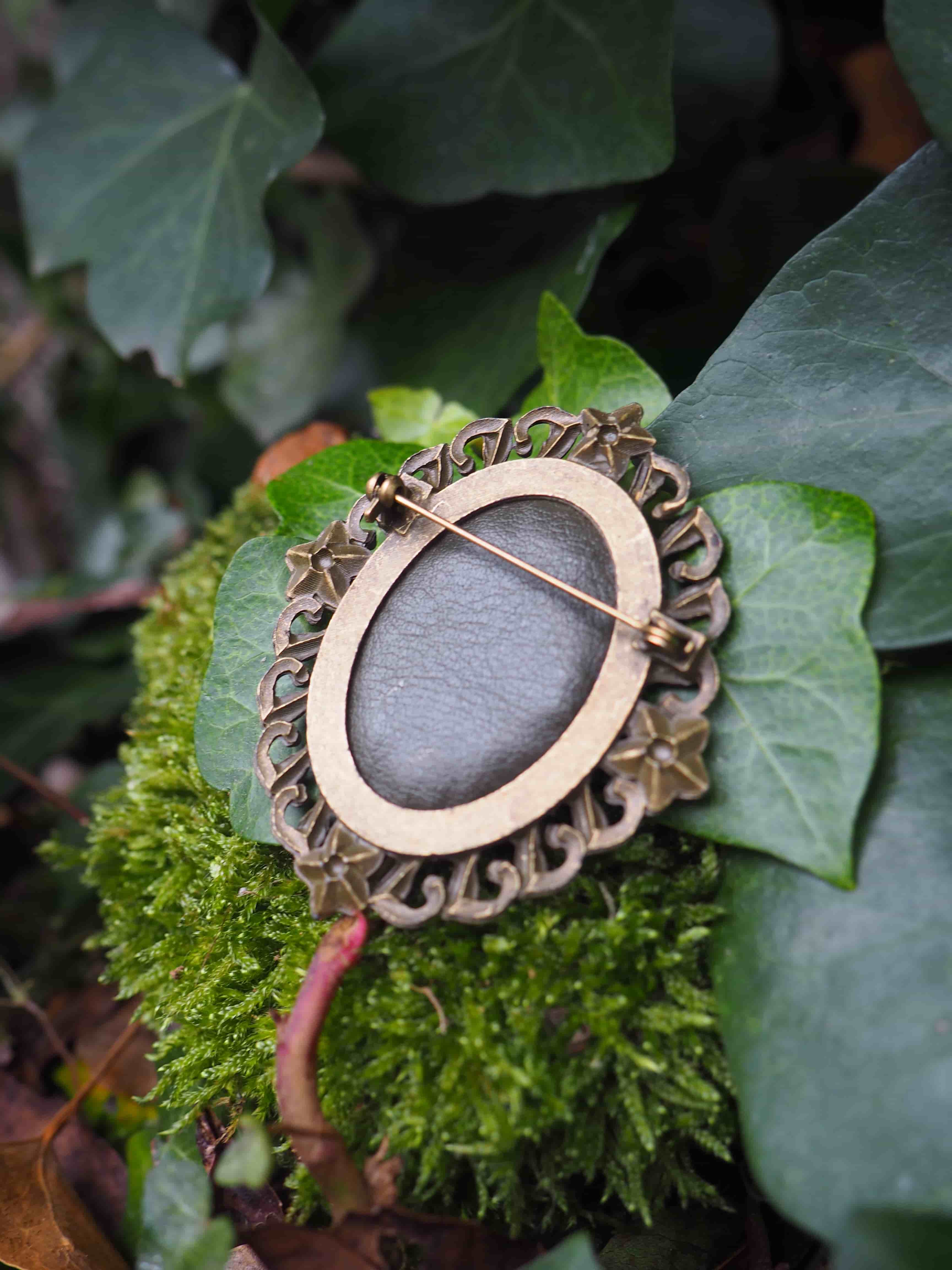 Cette image présente une broche ou un pendentif de style vintage posé délicatement sur un lit de mousse verte et de feuillage. Le bijou a une forme ovale avec un cadre décoratif en métal doré ou bronze, orné de motifs sculptés ou gravés qui lui donnent un aspect élégant et raffiné.  Au centre de la pièce se trouve une pierre ou un cabochon de couleur gris-violet, lisse et poli, qui contraste harmonieusement avec le métal doré du cadre. Une épingle métallique traverse le bijou, confirmant qu'il s'agit d'une broche fonctionnelle. L'ensemble repose sur un écrin naturel composé de mousse d'un vert tendre et de diverses feuilles aux nuances vertes variées, créant un arrière-plan organique qui met en valeur l'objet. Cette mise en scène évoque un style romantique ou bohème, où la nature sert d'écrin à ce bijou au charme rétro. L'éclairage doux et naturel de la photographie accentue les textures et les couleurs, créant une atmosphère poétique et intemporelle.