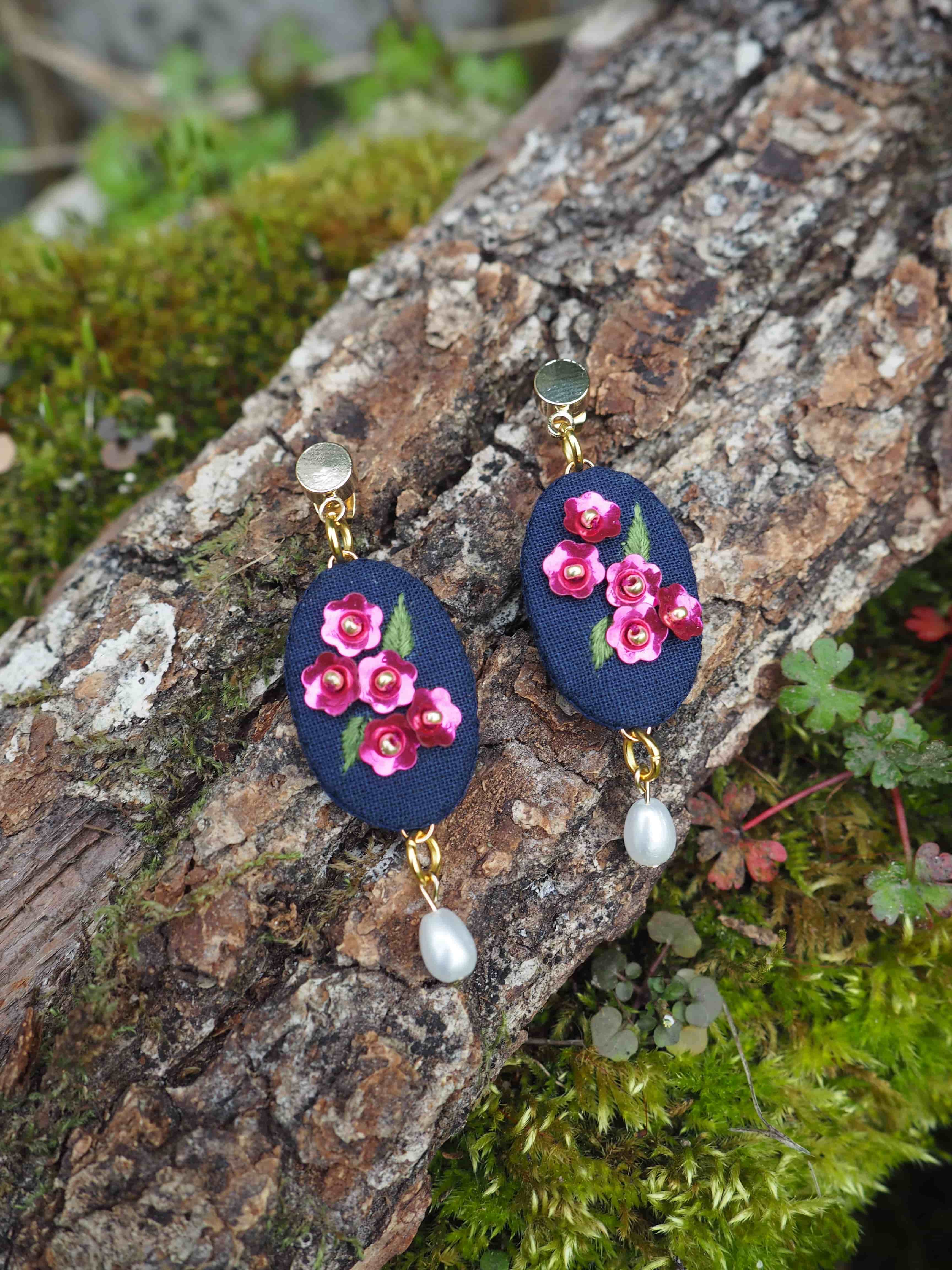 Cette image montre une paire de boucles d'oreilles artisanales posées sur un tronc d'arbre couvert de mousse. Les boucles d'oreilles sont composées d'un support en tissu bleu marine sur lequel sont brodées de délicates roses roses. Au bas de chaque boucle d'oreille pend une petite perle blanche. Les attaches semblent être en métal doré. La composition est très naturelle et romantique, avec le contraste entre les bijoux délicats et la texture rugueuse de l'écorce, ainsi que la mousse verte qui ajoute une touche de nature à l'image. Ces boucles d'oreilles ont un style vintage et fait-main très charmant.
