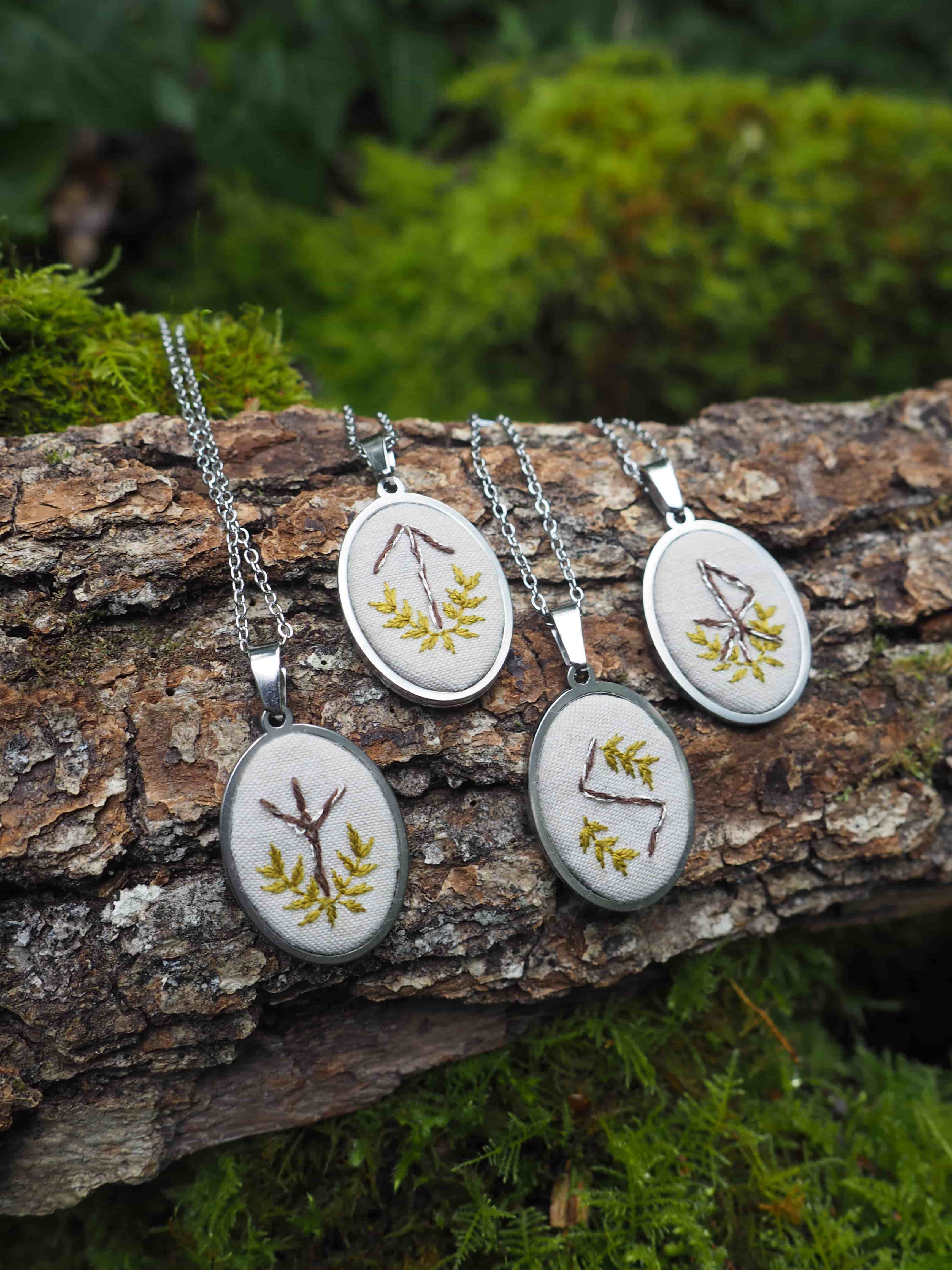L'image présente quatre pendentifs ovales, chacun orné d'un motif brodé, posés sur un morceau de bois recouvert de mousse. Voici une description détaillée des éléments visuels : *   **Les Pendentifs :**
    *   Il y a quatre pendentifs, tous de forme ovale et de taille similaire.
    *   Chaque pendentif est suspendu à une chaîne fine de couleur argentée, suggérant un matériau comme l'acier inoxydable ou l'argent.
    *   Le cadre de chaque pendentif est également de couleur argentée, avec un aspect métallique.
    *   À l'intérieur de chaque cadre, il y a un fond de tissu clair, probablement blanc cassé ou crème, sur lequel sont brodés des motifs.
    *   Les motifs brodés sont des symboles stylisés, qui ressemblent à des runes ou des caractères anciens, souvent accompagnés de petites branches ou feuilles stylisées. La broderie est réalisée avec du fil de couleur jaune-vert ou doré, créant un contraste doux avec le fond clair.
    *   Les pendentifs sont disposés en ligne sur le bois, avec deux à l'avant et deux légèrement en arrière. *   **Le Support (Bois et Mousse) :**
    *   Les pendentifs reposent sur un morceau de bois brut, qui semble être une branche ou un tronc d'arbre tombé. L'écorce du bois est visible, avec des textures et des crevasses naturelles.
    *   Le bois est partiellement recouvert de mousse verte, ajoutant une touche naturelle et organique à la scène. La mousse est d'un vert vif et luxuriant par endroits. *   **L'Arrière-plan :**
    *   L'arrière-plan est flou (bokeh), ce qui met l'accent sur les pendentifs au premier plan.
    *   On distingue des teintes de vert foncé et de brun, suggérant un environnement forestier ou boisé, avec d'autres éléments végétaux et de la mousse. *   **L'Ambiance Générale :**
    *   L'image dégage une atmosphère naturelle, artisanale et un peu mystique, en raison des symboles brodés et du cadre forestier.
    *   La lumière semble douce et diffuse, typique d'un sous-bois. En résumé, l'image met en scène des bijoux artisanaux (pendentifs brodés) dans un cadre naturel et rustique, soulignant leur caractère unique et potentiellement inspiré par la nature ou des traditions anciennes.