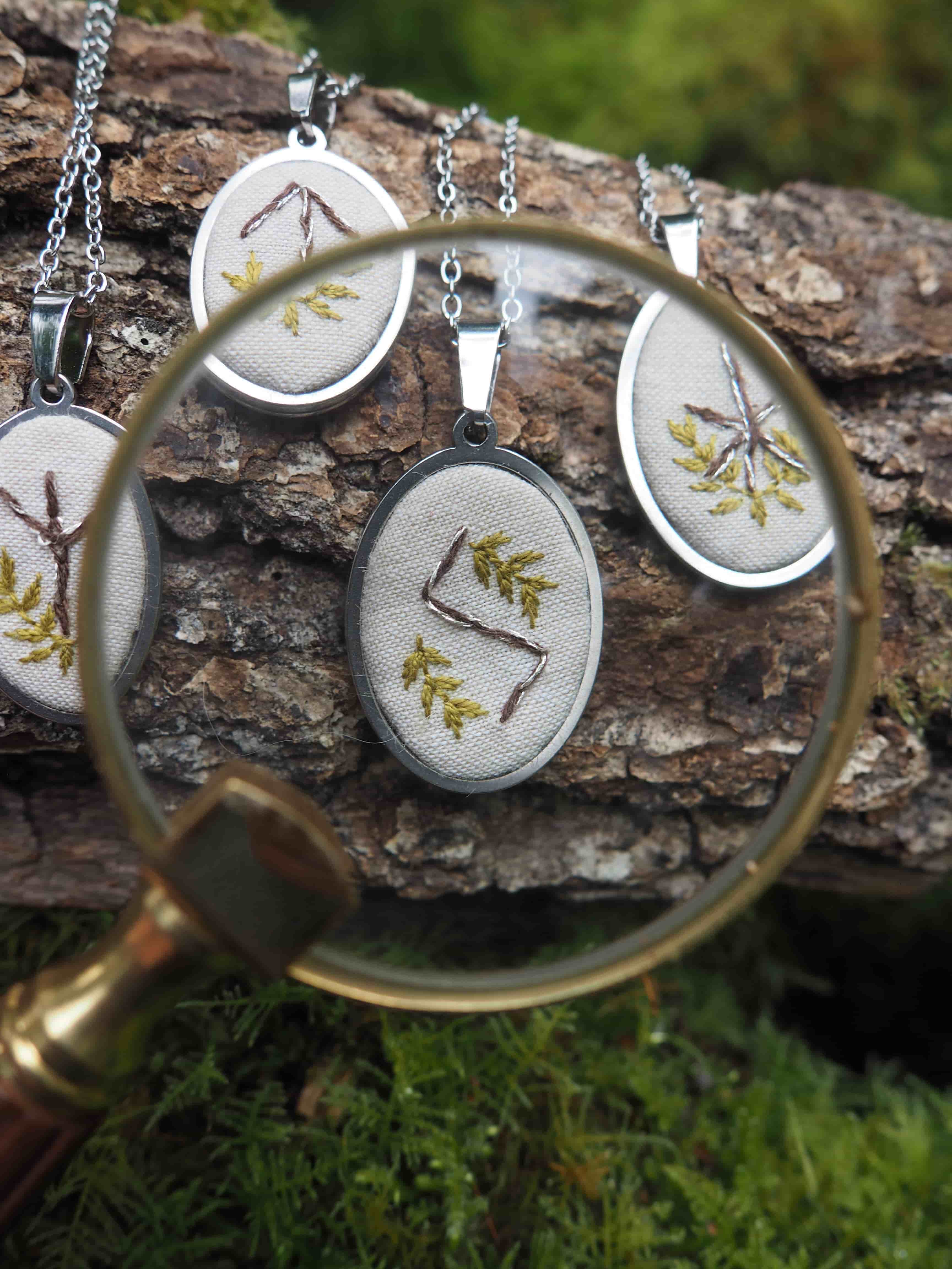 Cette image présente une composition artistique et détaillée de plusieurs pendentifs brodés, mis en valeur par une loupe. Au premier plan, une loupe à monture en laiton doré et manche en bois est positionnée de manière à encadrer l'un des pendentifs. La lentille de la loupe offre une vue agrandie et nette du pendentif qu'elle couvre, révélant les détails de la broderie. Les pendentifs sont de forme ovale, avec un cadre métallique argenté brillant. Chaque pendentif contient une petite broderie sur un fond de tissu clair, probablement du lin ou du coton écru. Les broderies représentent des motifs stylisés, qui ressemblent à des runes ou des symboles anciens, souvent accompagnés de petites branches ou feuilles brodées en vert et jaune, évoquant des éléments naturels. Les pendentifs sont suspendus à des chaînes fines et argentées. Ils sont disposés sur une surface texturée qui semble être un morceau d'écorce d'arbre ou une branche recouverte de mousse verte, ce qui renforce l'ambiance naturelle et rustique de la scène. La mousse est particulièrement visible sous la loupe et autour des pendentifs, ajoutant de la profondeur et de la couleur à l'arrière-plan. L'éclairage est doux et naturel, mettant en évidence les textures des matériaux et les couleurs des broderies. La mise au point est sélective, avec la loupe et le pendentif qu'elle encadre étant les plus nets, tandis que les autres pendentifs et l'arrière-plan sont légèrement flous, créant une profondeur de champ agréable. L'ensemble de l'image dégage une atmosphère artisanale, mystique et connectée à la nature, suggérant que ces pendentifs pourraient être des talismans ou des objets symboliques.