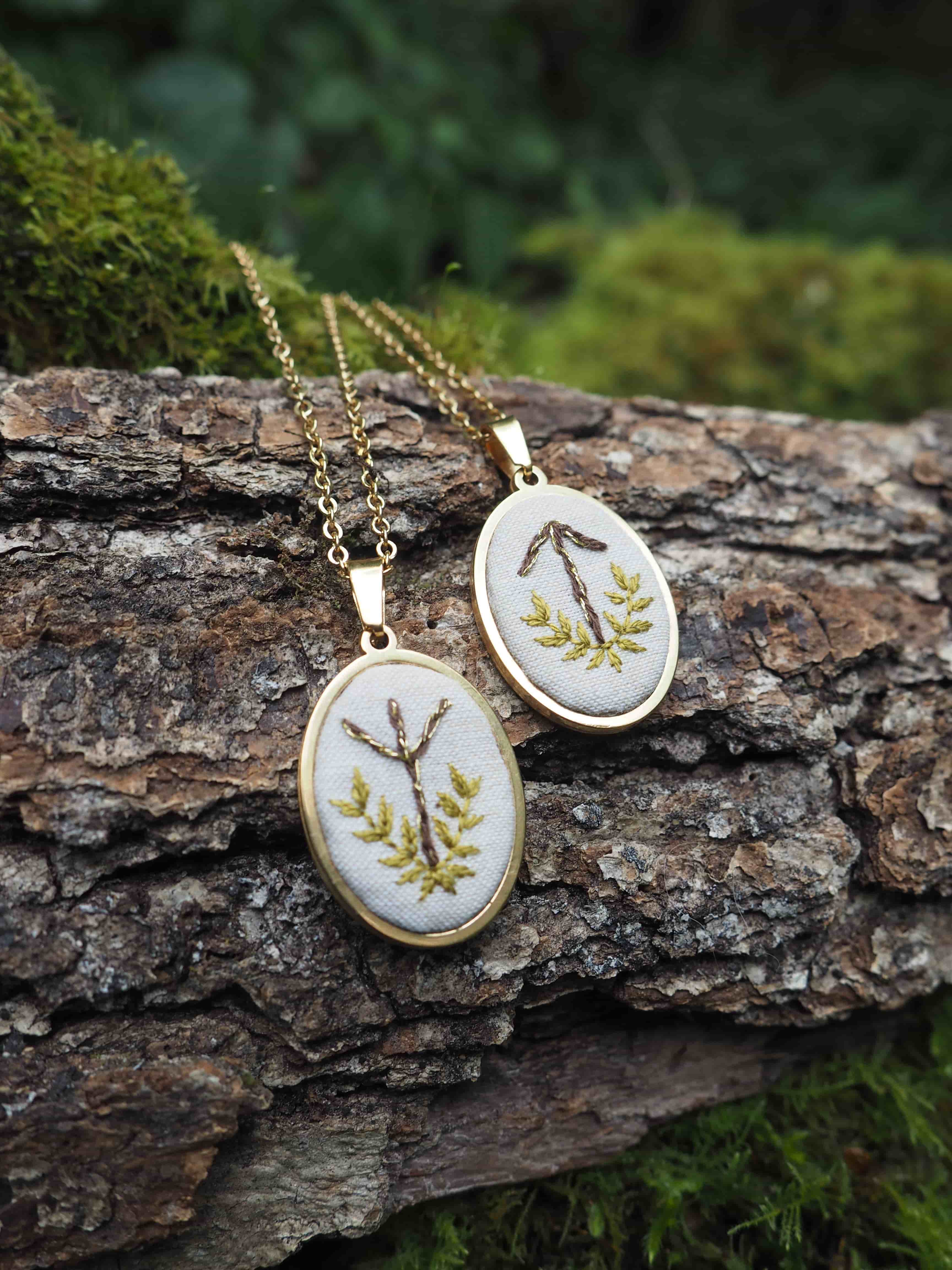 Cette image présente deux pendentifs ovales en laiton doré, chacun suspendu à une fine chaîne dorée. Les pendentifs sont posés sur un morceau de bois brut, probablement une bûche ou une branche, dont l'écorce est texturée et de couleur marron foncé. Chaque pendentif contient une broderie sur un fond de tissu clair, probablement blanc cassé ou crème. Les broderies représentent des motifs végétaux stylisés, évoquant des branches ou des feuilles.
Le pendentif de gauche montre un motif qui ressemble à une branche d'arbre avec plusieurs ramifications, brodée en fil marron et agrémentée de petites feuilles vertes ou dorées.
Le pendentif de droite présente un motif similaire, mais la forme principale est plus anguleuse, rappelant une ancre ou une lettre stylisée, également brodée en fil marron avec des feuilles vertes ou dorées de chaque côté de la base. L'arrière-plan est flou, mais on distingue des tons de vert et de marron, suggérant un environnement naturel, peut-être une forêt ou un jardin, avec de la mousse et de la végétation. La lumière semble naturelle et douce, mettant en valeur les détails des pendentifs. L'ensemble de l'image dégage une atmosphère artisanale et naturelle, soulignant la beauté des bijoux faits à la main et leur lien avec la nature.