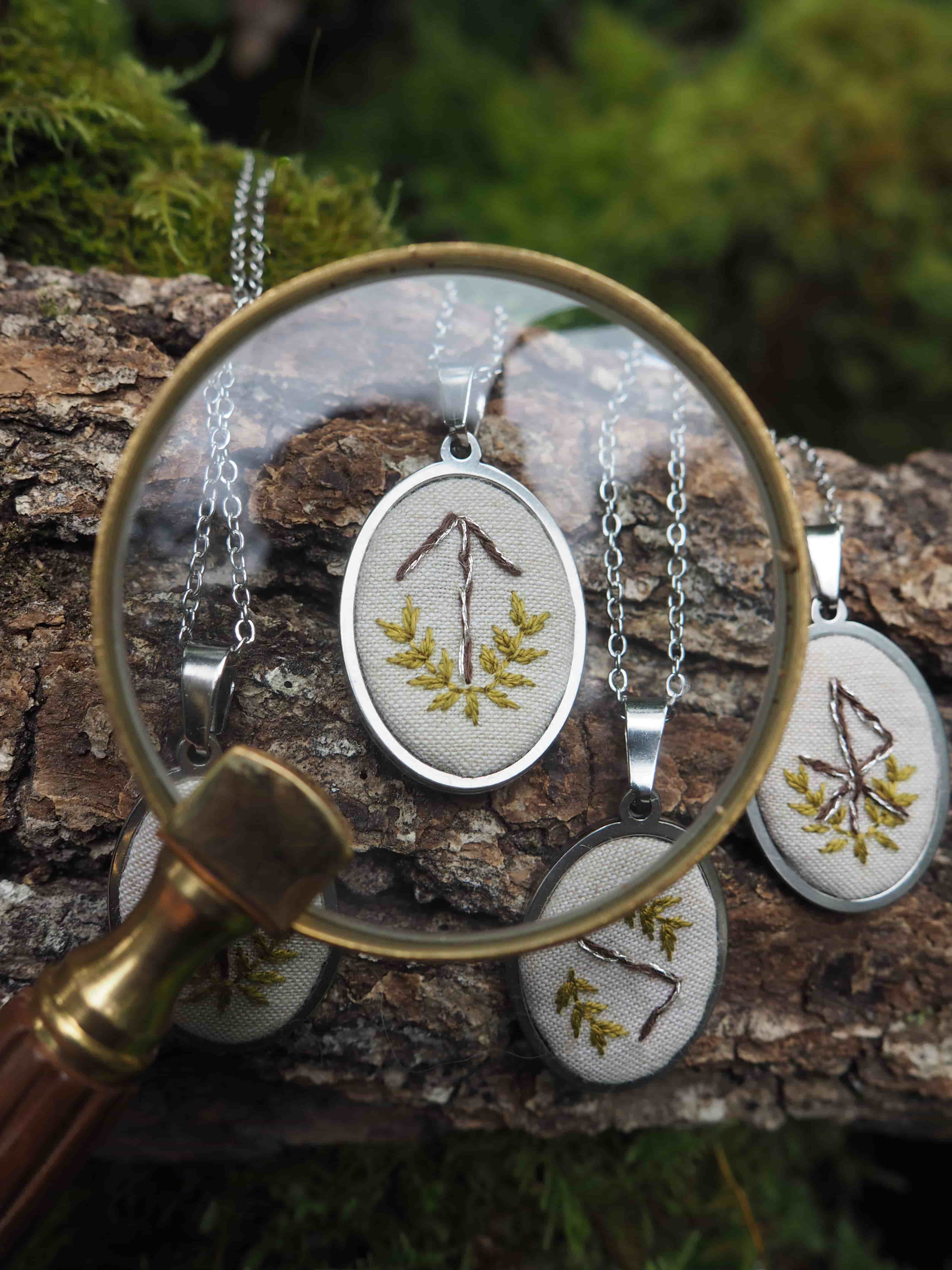 Cette image présente une composition artistique et naturelle, centrée sur des pendentifs brodés, observés à travers une loupe. Au premier plan, une loupe à monture en laiton et manche en bois est positionnée de manière à agrandir l'un des pendentifs. La lentille de la loupe est claire et montre une broderie détaillée d'un motif qui ressemble à une rune ou un symbole stylisé, composé de branches marron et de feuilles vertes, sur un fond de tissu clair, probablement du lin ou du coton. Le pendentif est de forme ovale, encadré d'un métal argenté et suspendu à une fine chaîne argentée. Autour de la loupe et en arrière-plan, plusieurs autres pendentifs similaires sont visibles, certains partiellement cachés par la loupe. Ils sont tous de forme ovale, avec des broderies de motifs végétaux ou runiques variés, et sont également suspendus à des chaînes. L'ensemble des pendentifs repose sur un morceau de bois brut, probablement une branche ou un tronc d'arbre, dont l'écorce est texturée et de couleur marron-gris. En arrière-plan, on distingue de la mousse verte et d'autres éléments végétaux flous, suggérant un environnement extérieur, peut-être une forêt ou un jardin. La lumière semble naturelle et douce, mettant en valeur les textures des broderies, du bois et de la loupe. L'ambiance générale est artisanale, naturelle et un peu mystique, évoquant un travail manuel minutieux et une connexion avec la nature.