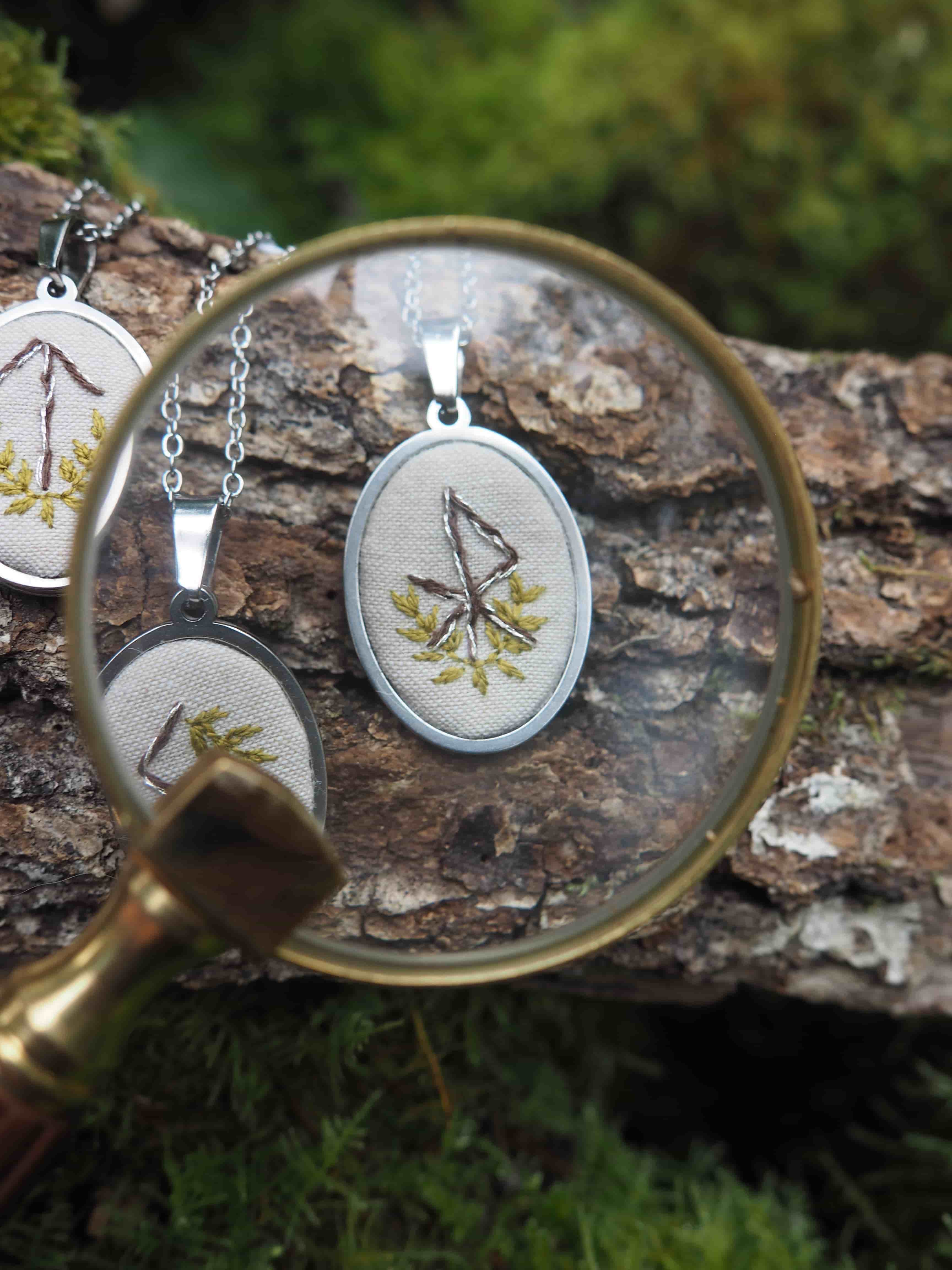L'image présente une composition artistique et détaillée, centrée sur un pendentif brodé, observé à travers une loupe. Au premier plan, une loupe à monture dorée et manche en bois est tenue, son verre grossissant une partie de la scène. À travers la loupe, on peut voir un pendentif ovale. Ce pendentif est composé d'un cadre métallique argenté qui entoure un tissu de couleur crème ou beige clair. Sur ce tissu, une broderie représente un symbole qui ressemble à une rune ou un caractère stylisé, réalisé avec du fil de couleur marron ou ocre. Autour de ce symbole, de petites feuilles ou branches stylisées sont brodées en fil vert, ajoutant une touche naturelle. En arrière-plan, hors du champ de la loupe mais toujours visible, se trouvent d'autres pendentifs similaires, suggérant une collection ou une série. L'un d'eux est partiellement visible à gauche, montrant un autre symbole brodé. Tous les pendentifs sont suspendus à des chaînes fines et argentées. Les pendentifs reposent sur un morceau de bois brut, probablement une écorce ou une branche, dont la texture est bien visible avec ses crevasses et ses motifs naturels. Le bois est de couleur marron foncé. L'arrière-plan plus lointain est flou, mais on distingue des teintes de vert, suggérant un environnement naturel comme de la mousse ou de la végétation, ce qui renforce l'aspect organique et artisanal de la scène. L'éclairage semble naturel, mettant en valeur les textures et les couleurs des objets. L'ensemble de l'image dégage une atmosphère de minutie, d'artisanat et de connexion avec la nature.