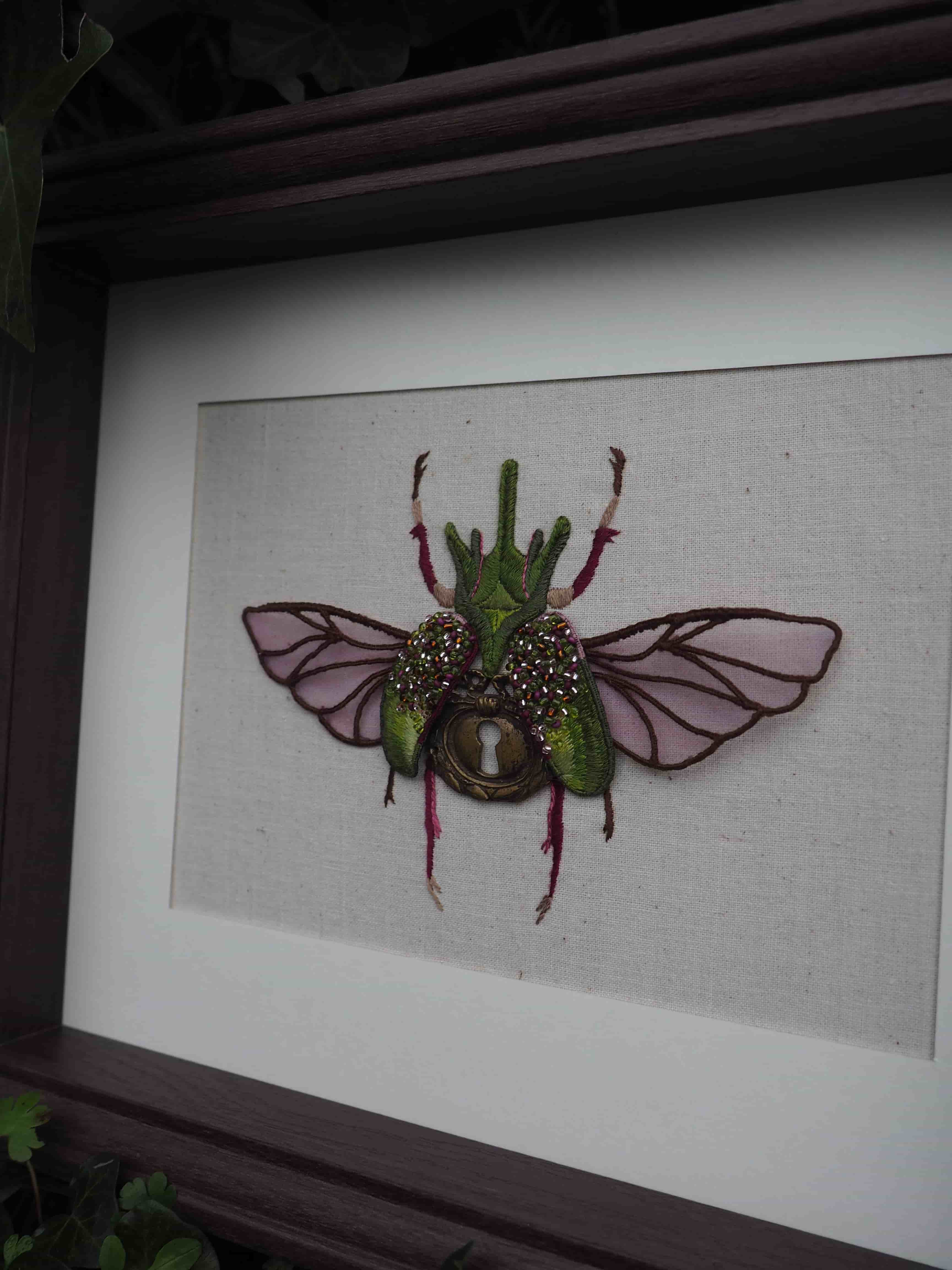Cette image montre une œuvre d'art encadrée représentant un insecte décoratif. Il s'agit d'une création artistique d'un scarabée ou d'un coléoptère avec des ailes déployées, réalisée dans un style mixte combinant différents matériaux. L'insecte est de couleur verte avec des ailes translucides ou rosées, et semble avoir des détails brillants ou métalliques. Il est présenté sur un fond clair, probablement en tissu ou en papier, dans un cadre sombre. C'est une pièce décorative qui mélange l'aspect naturaliste des insectes avec une touche artistique et fantaisiste.