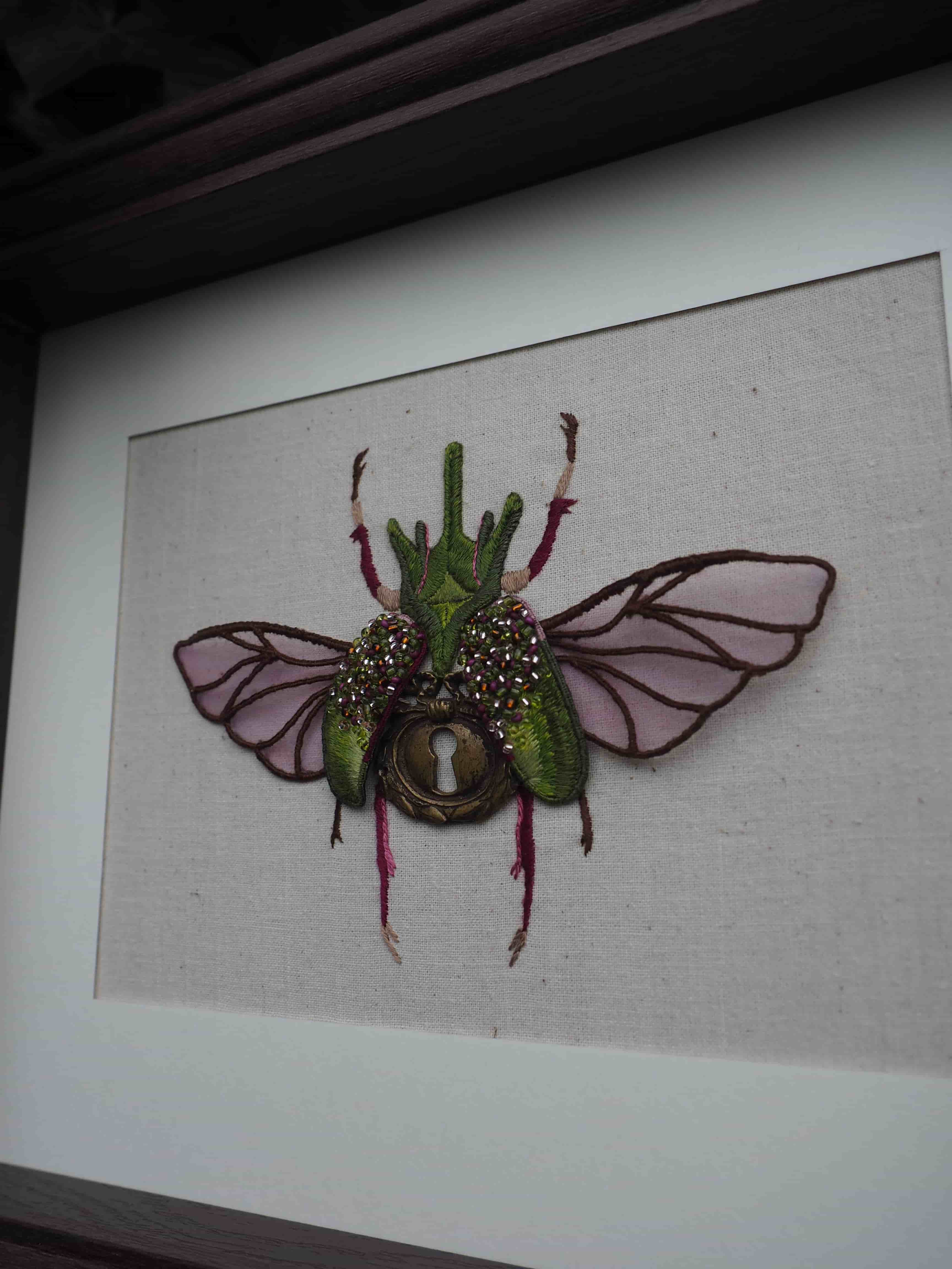 Cette image montre une œuvre d'art textile encadrée représentant un scarabée ou un insecte stylisé. L'insecte est créé avec différents matériaux et textures, notamment du tissu vert pour le corps, des ailes transparentes ou violettes, et ce qui semble être des éléments décoratifs brillants ou pailletés. Au centre de l'insecte se trouve ce qui ressemble à un œil ou une pierre décorative. L'œuvre est présentée sur un fond clair, probablement en lin ou en toile, et est encadrée dans ce qui semble être un cadre sombre. C'est une pièce décorative qui mélange l'art textile et l'entomologie artistique, créant un effet à la fois naturaliste et fantaisiste.