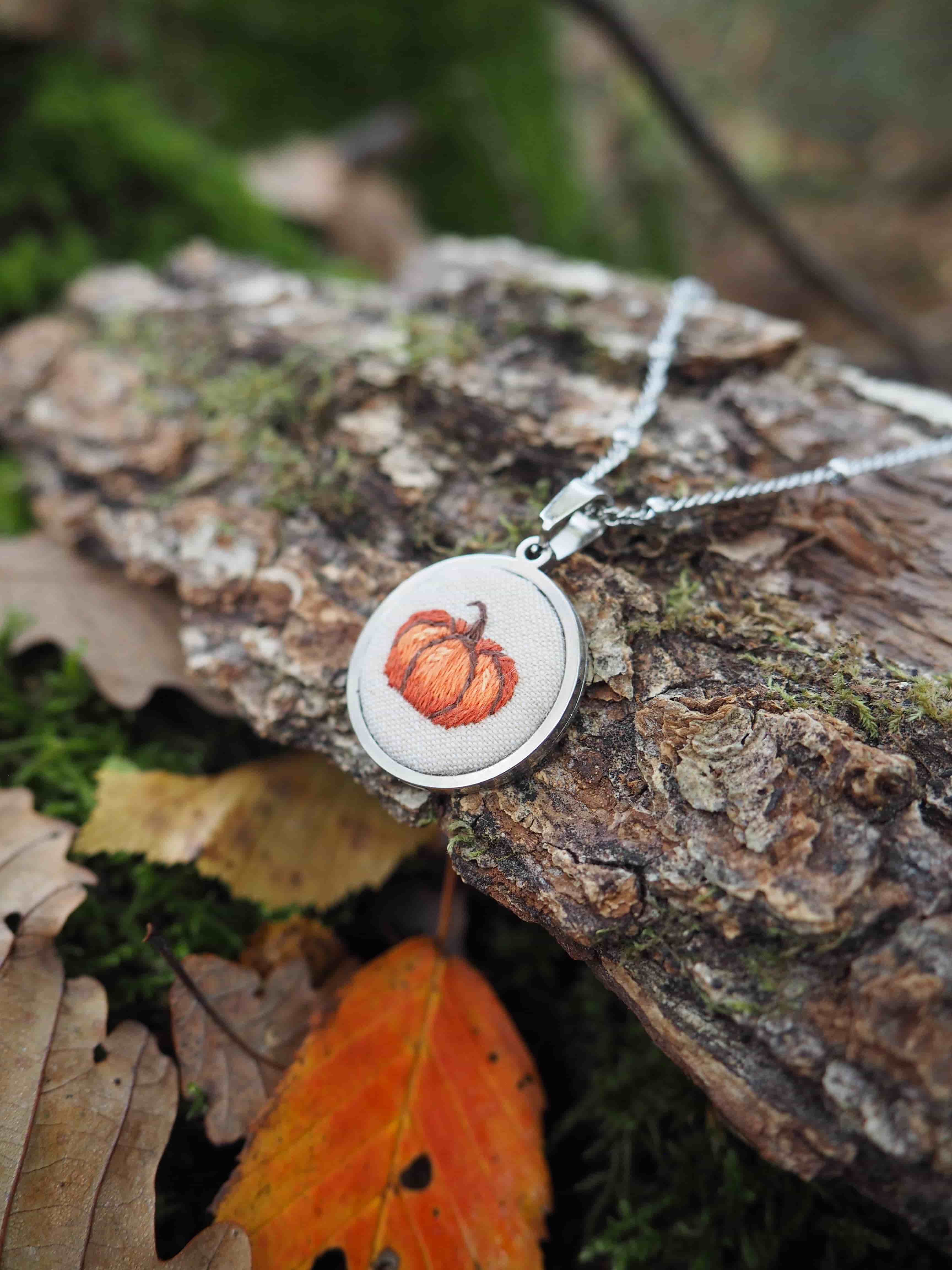 Cette image présente un bijou délicat posé dans un environnement naturel automnal. Au centre de la composition se trouve un pendentif circulaire en métal argenté, suspendu à une chaîne fine. Le pendentif contient un motif décoratif représentant une citrouille orange, évoquant l'esprit de l'automne et d'Halloween. Le bijou repose sur un morceau d'écorce rugueuse et texturée, probablement d'un arbre, qui présente des tons bruns et gris naturels avec quelques traces de mousse verte. L'arrière-plan est artistiquement flou, créant un effet de profondeur de champ qui met en valeur le pendentif. L'environnement est typiquement automnal, avec des feuilles mortes visibles - notamment une feuille orange-rougeâtre au premier plan et d'autres feuilles brunâtres dispersées autour. La végétation verte floue en arrière-plan suggère un cadre forestier ou de jardin. L'éclairage naturel et doux crée une atmosphère chaleureuse et organique, parfaitement adaptée pour mettre en valeur ce bijou à thème saisonnier. La composition photographique équilibre harmonieusement les éléments naturels avec l'objet artisanal.