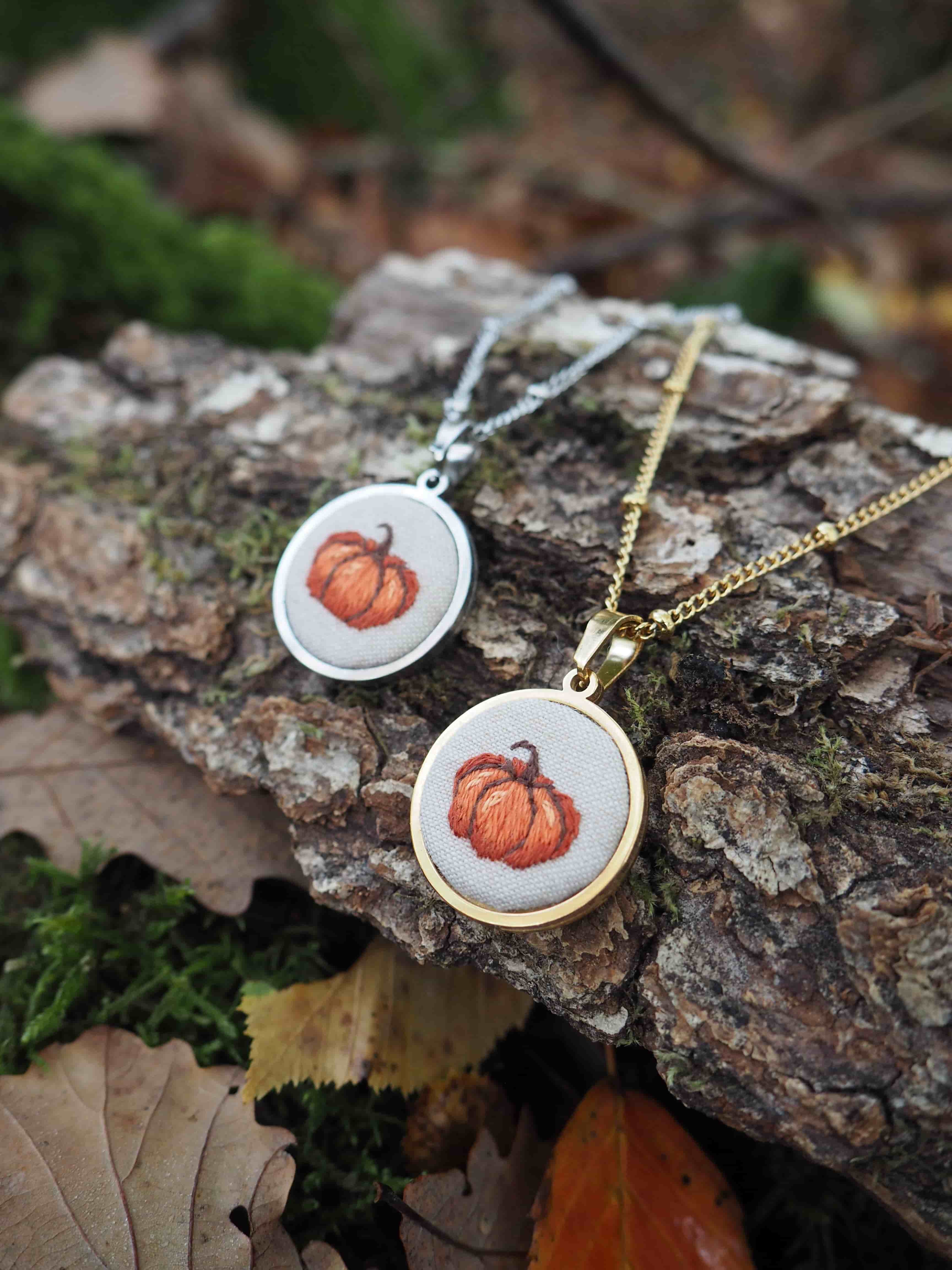 Cette image présente deux colliers pendentifs disposés sur un morceau d'écorce dans un environnement naturel automnal.  **Les bijoux :**
- Deux pendentifs circulaires identiques avec des montures dorées
- Chacun contient une broderie délicate représentant une citrouille orange
- Un collier a une chaîne argentée, l'autre une chaîne dorée
- Le travail de broderie est fin et détaillé, donnant un aspect artisanal **Le décor :**
- Les pendentifs reposent sur un morceau d'écorce texturée
- L'arrière-plan présente des feuilles mortes dans les tons bruns et dorés
- On aperçoit de la mousse verte qui ajoute une touche de couleur naturelle
- L'ensemble évoque parfaitement l'atmosphère automnale **L'ambiance :**
Cette mise en scène photographique met en valeur ces bijoux à thème saisonnier, parfaits pour célébrer l'automne ou Halloween. La présentation sur des éléments naturels renforce le côté authentique et fait-main de ces accessoires.