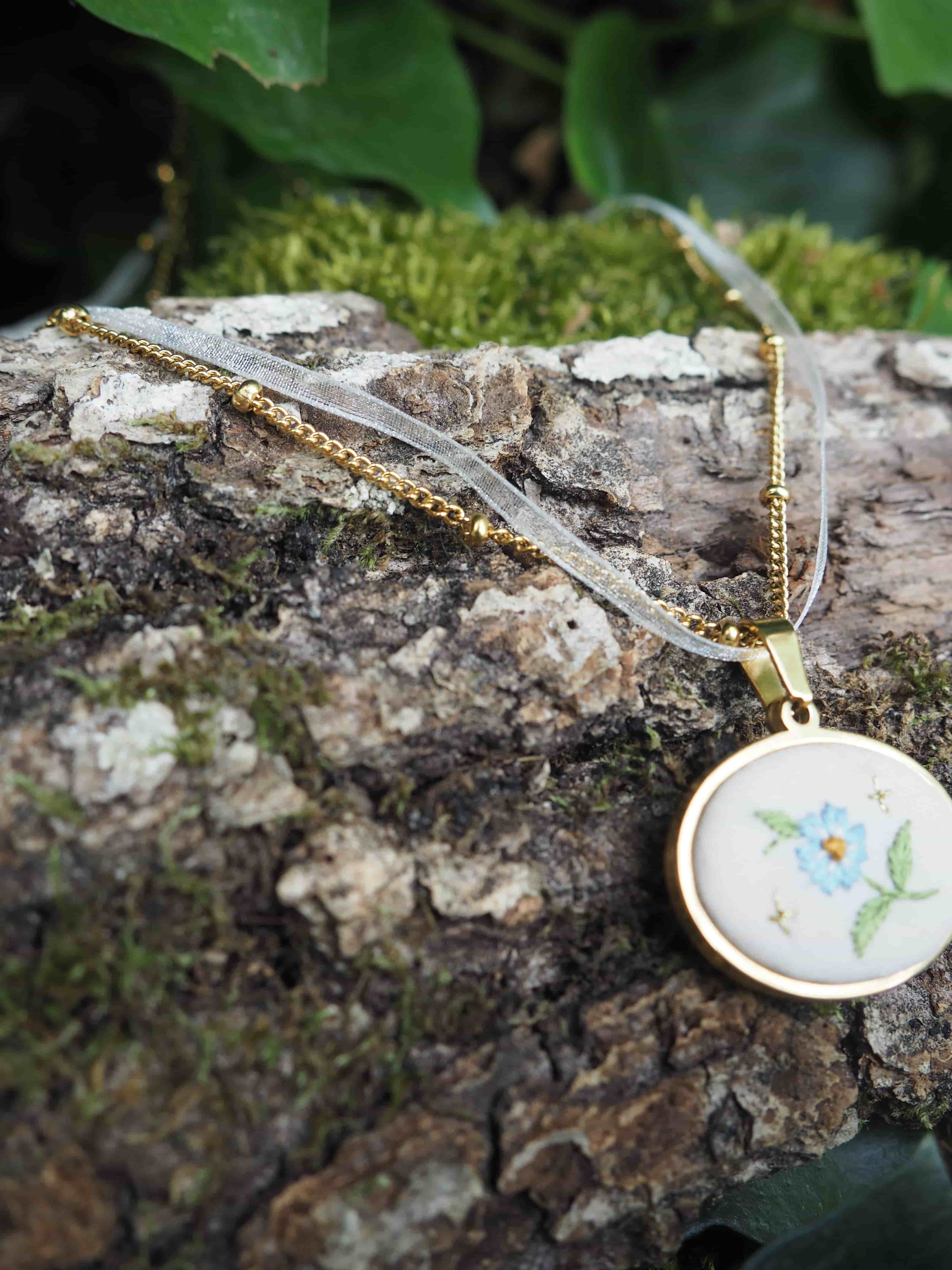 Cette image montre un délicat pendentif sur une chaîne dorée, posé sur un morceau d'écorce d'arbre couvert de mousse. Le pendentif est de forme ovale avec un fond blanc et comporte une délicate broderie florale, on peut distinguer une petite fleur bleue avec des feuilles vertes. La composition est très naturelle et poétique, avec le contraste entre le bijou raffiné et la texture rugueuse de l'écorce, ainsi que la verdure de la mousse en arrière-plan. C'est une mise en scène qui évoque un style romantique et bucolique.