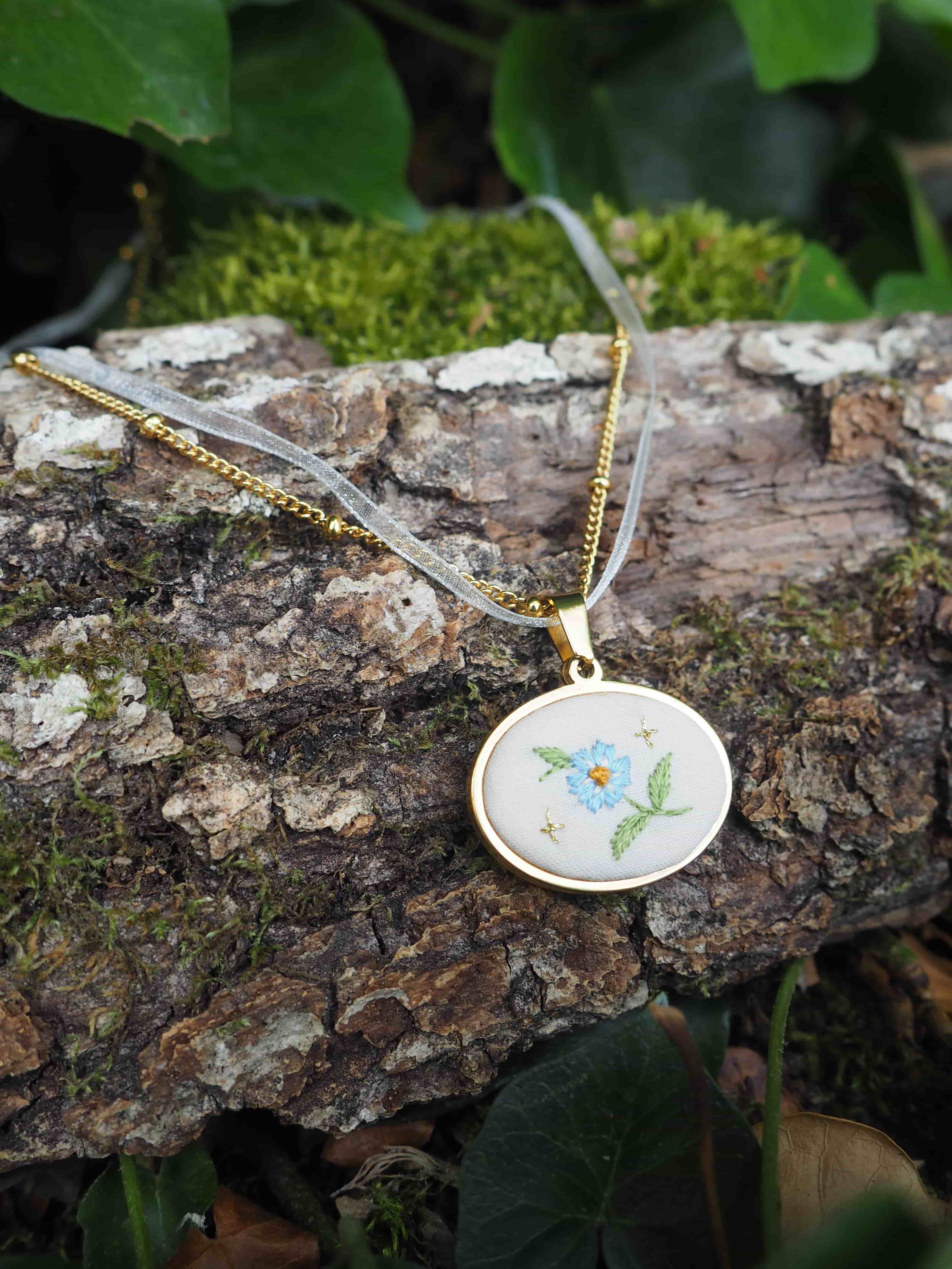 L'image montre un délicat pendentif brodé posé sur un morceau d'écorce d'arbre couvert de mousse. Le pendentif est de forme ronde, monté dans un cadre doré, et suspendu à une fine chaîne également dorée. La broderie sur le pendentif représente un motif floral avec ce qui semble être une petite fleur bleue et des feuilles vertes sur un fond blanc. L'arrière-plan naturel avec l'écorce texturée, la mousse verte et quelques feuilles crée une belle mise en scène naturelle qui met en valeur ce bijou artisanal délicat.