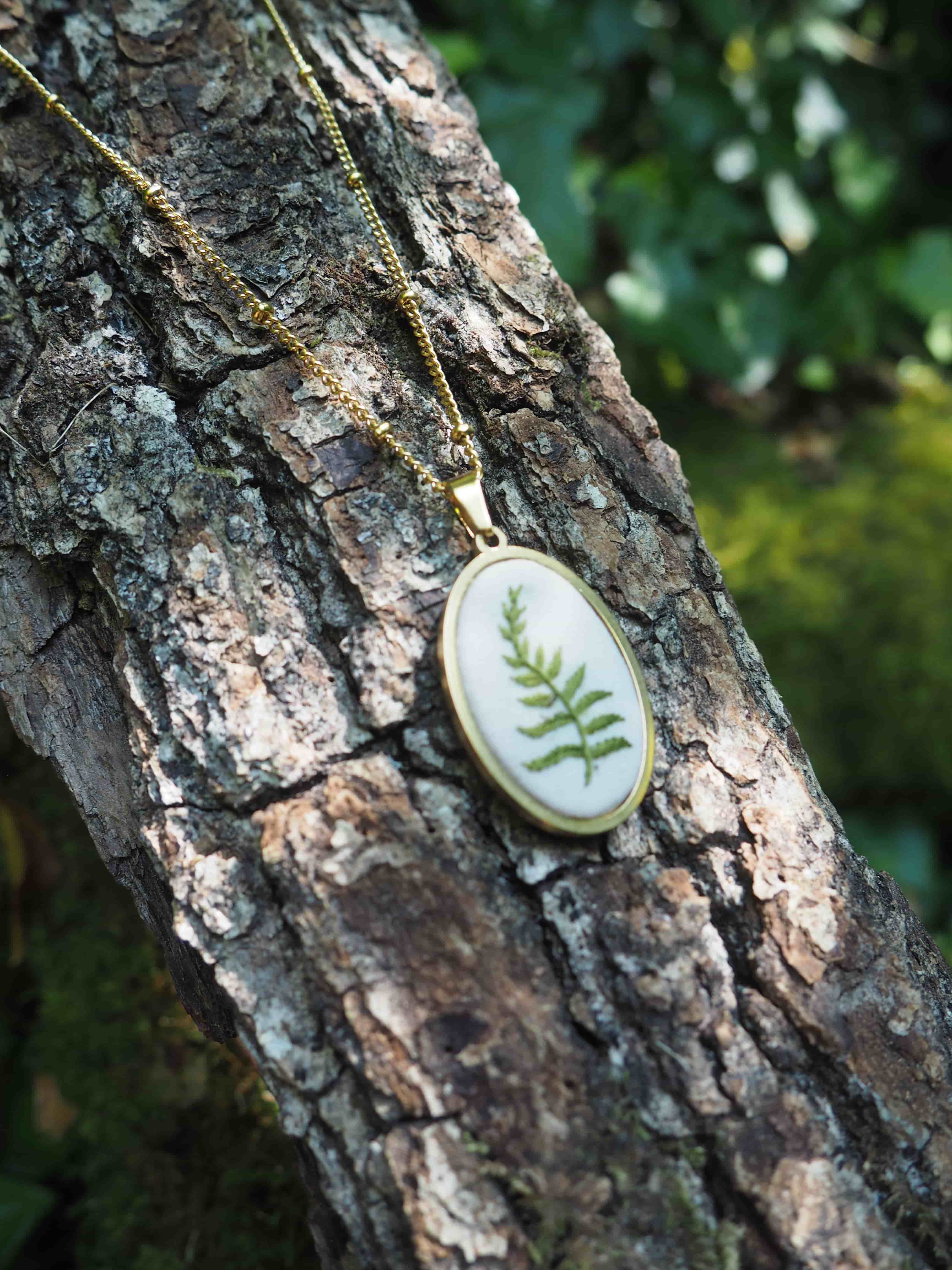L'image montre des pendentifs ou des bijoux délicats qui semblent être faits avec des feuilles de fougère préservées dans ce qui paraît être de la résine ou du verre, montés sur des chaînes dorées. Les pendentifs sont photographiés sur l'écorce rugueuse d'un arbre, créant un contraste intéressant entre la texture naturelle brute de l'écorce et la finesse des bijoux. La fougère verte à l'intérieur des pendentifs circulaires blancs crée un effet botanique élégant et naturel. L'arrière-plan est légèrement flou mais on peut apercevoir de la verdure, ce qui complète bien la thématique naturelle de la composition.