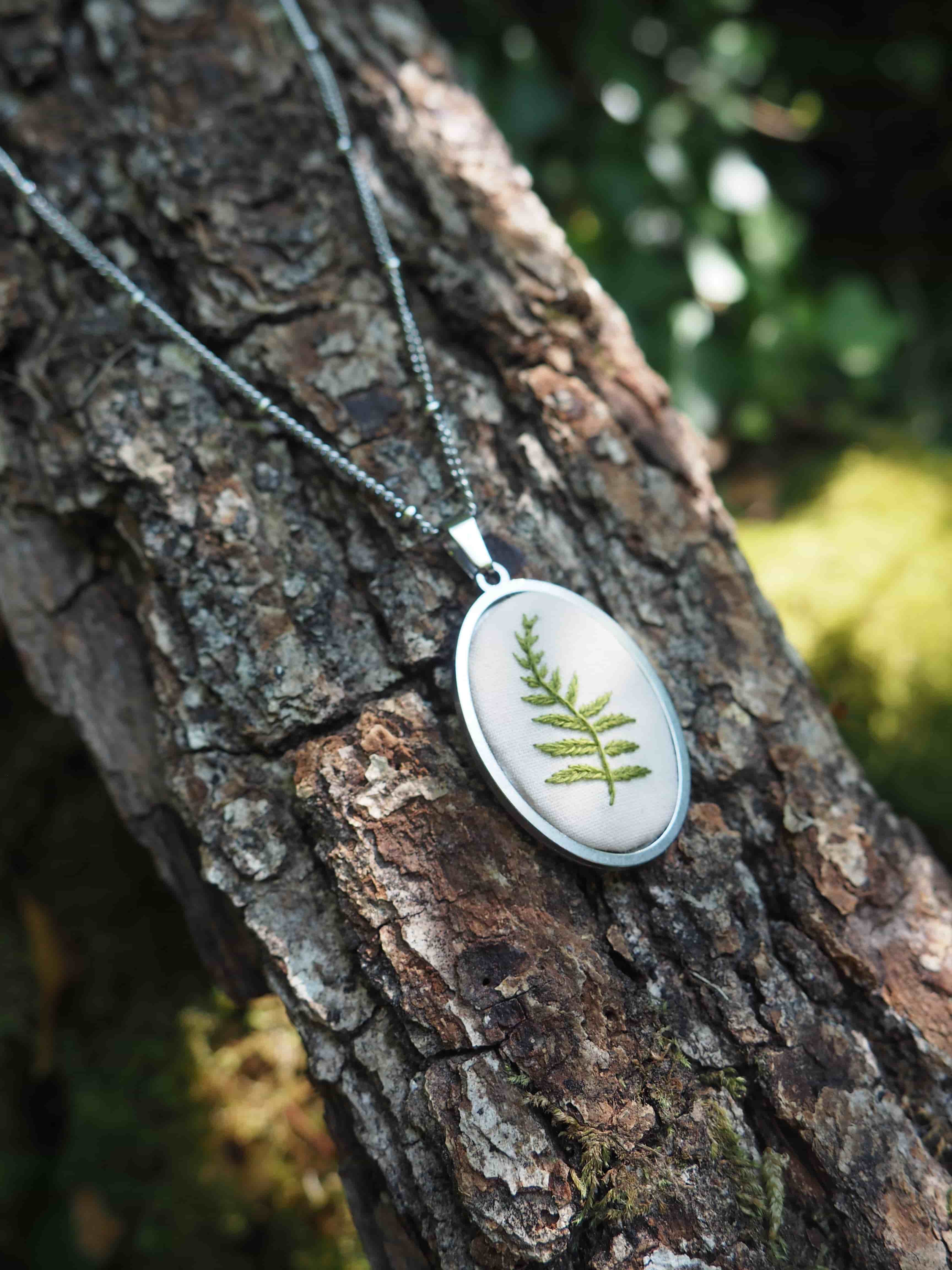 Cette image montre un délicat pendentif posé sur l'écorce d'un arbre. Le pendentif est de forme ovale avec un cadre en argent, et contient une véritable feuille de fougère préservée sous ce qui semble être une résine transparente. Il est attaché à une fine chaîne argentée. La photo est prise en gros plan avec un bel effet de profondeur de champ, l'arrière-plan verdoyant étant légèrement flou. L'écorce rugueuse et texturée de l'arbre crée un contraste intéressant avec la finesse et la délicatesse du bijou. C'est une belle composition qui met en valeur cette pièce de bijouterie naturelle et artisanale.