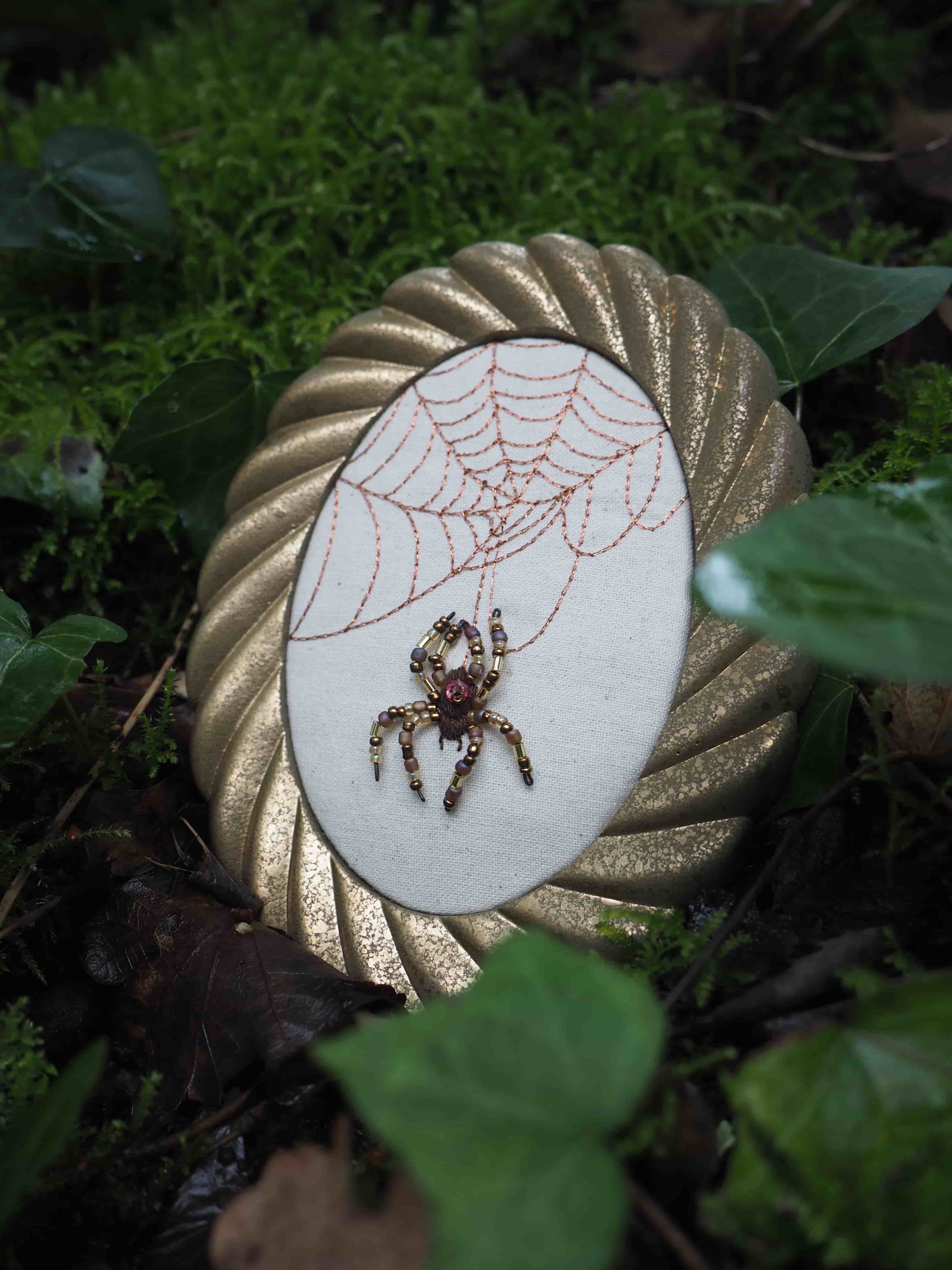 Cette image présente un cadre ovale décoratif posé sur un lit de mousse et de feuilles dans un environnement naturel, probablement une forêt ou un jardin. Le cadre est de couleur dorée, avec une texture plissée ou torsadée sur son pourtour, lui donnant un aspect riche et travaillé. À l'intérieur du cadre, sur un fond de tissu clair (peut-être du lin ou du coton écru), se trouve une broderie délicate. La broderie représente une toile d'araignée, réalisée avec un fil fin de couleur cuivrée ou marron clair, et une araignée. L'araignée est particulièrement détaillée, elle semble être faite de perles et de petits éléments brillants, peut-être des pierres ou des cristaux, de couleur marron foncé et rouge-violet, lui conférant un aspect tridimensionnel et scintillant. Elle est suspendue à la toile, comme si elle était en train de descendre. L'arrière-plan est composé de mousse verte luxuriante et de feuilles d'arbres, certaines étant des feuilles de lierre vert foncé. La lumière est douce, créant une atmosphère un peu mystérieuse et naturelle. L'ensemble de la scène évoque un sentiment d'artisanat, de nature et peut-être une touche de fantaisie ou de gothique doux.