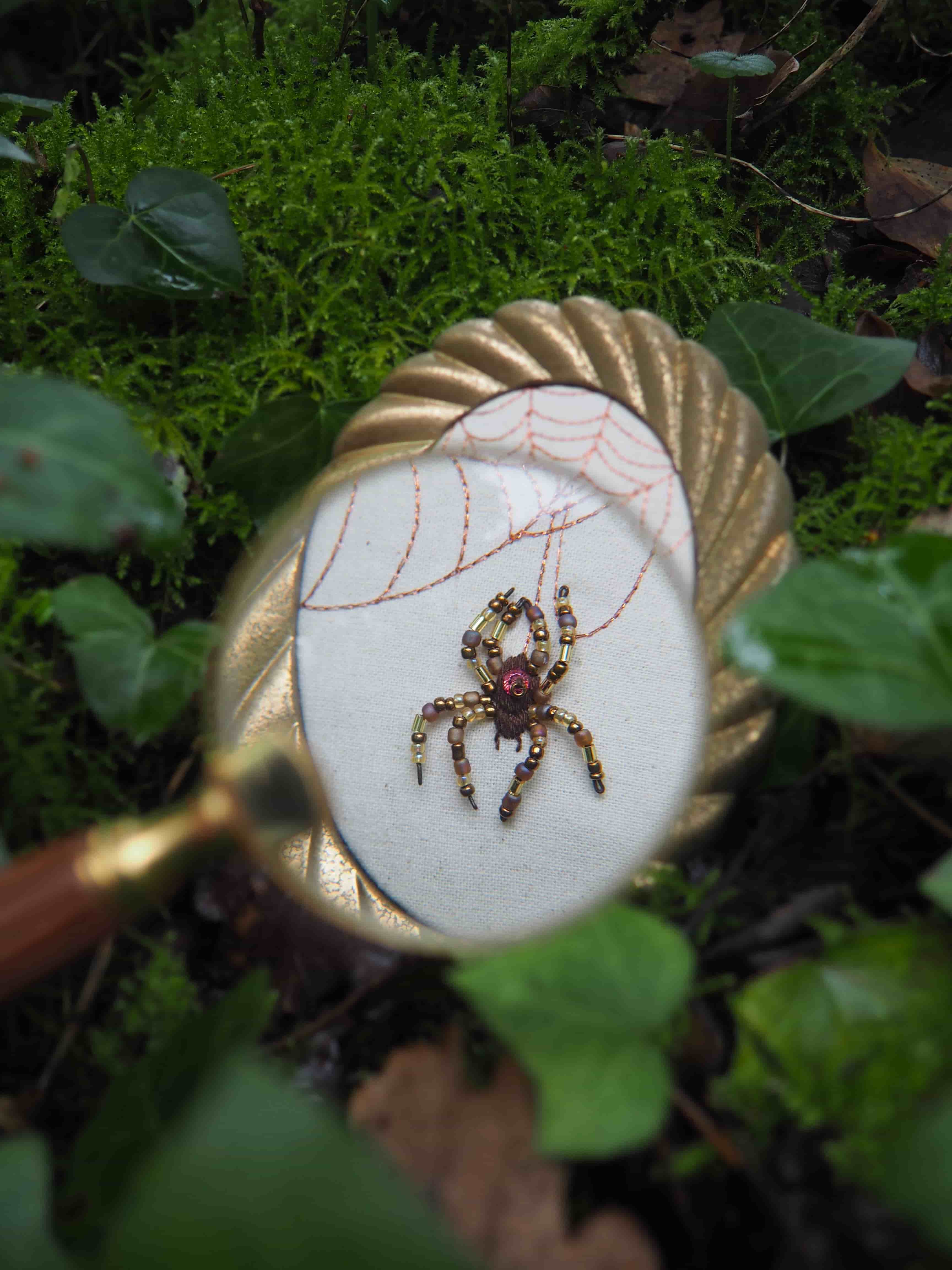 Cette image présente une composition intrigante et détaillée, centrée sur une loupe qui révèle un motif brodé. Au premier plan, on distingue des feuilles de lierre et de la mousse verte, créant un environnement naturel et légèrement flou qui encadre la scène. La loupe, avec son cadre doré et son manche en bois visible sur la gauche, est l'élément central. À travers la lentille de la loupe, on observe un motif brodé sur un tissu de couleur claire, probablement blanc cassé ou crème. Le motif représente une araignée détaillée, réalisée avec des perles de différentes couleurs : le corps est principalement composé de perles marron-doré, et une perle plus grande et de couleur rouge-violet orne le thorax de l'araignée, ajoutant une touche de couleur vive. L'araignée est suspendue à une toile d'araignée stylisée, brodée avec du fil de couleur rouille ou marron clair, qui s'étend sur le tissu. L'arrière-plan, au-delà de la loupe, est également constitué de mousse verte et de quelques feuilles, rappelant l'environnement du premier plan. La profondeur de champ est faible, ce qui met en évidence la loupe et son contenu, tandis que le reste de l'image est légèrement flou, créant un effet de mise au point sélective. L'éclairage semble naturel et diffus, mettant en valeur les textures de la mousse et les détails de la broderie. L'ensemble de l'image évoque une ambiance de découverte, de curiosité et d'appréciation des petits détails de la nature et de l'artisanat.