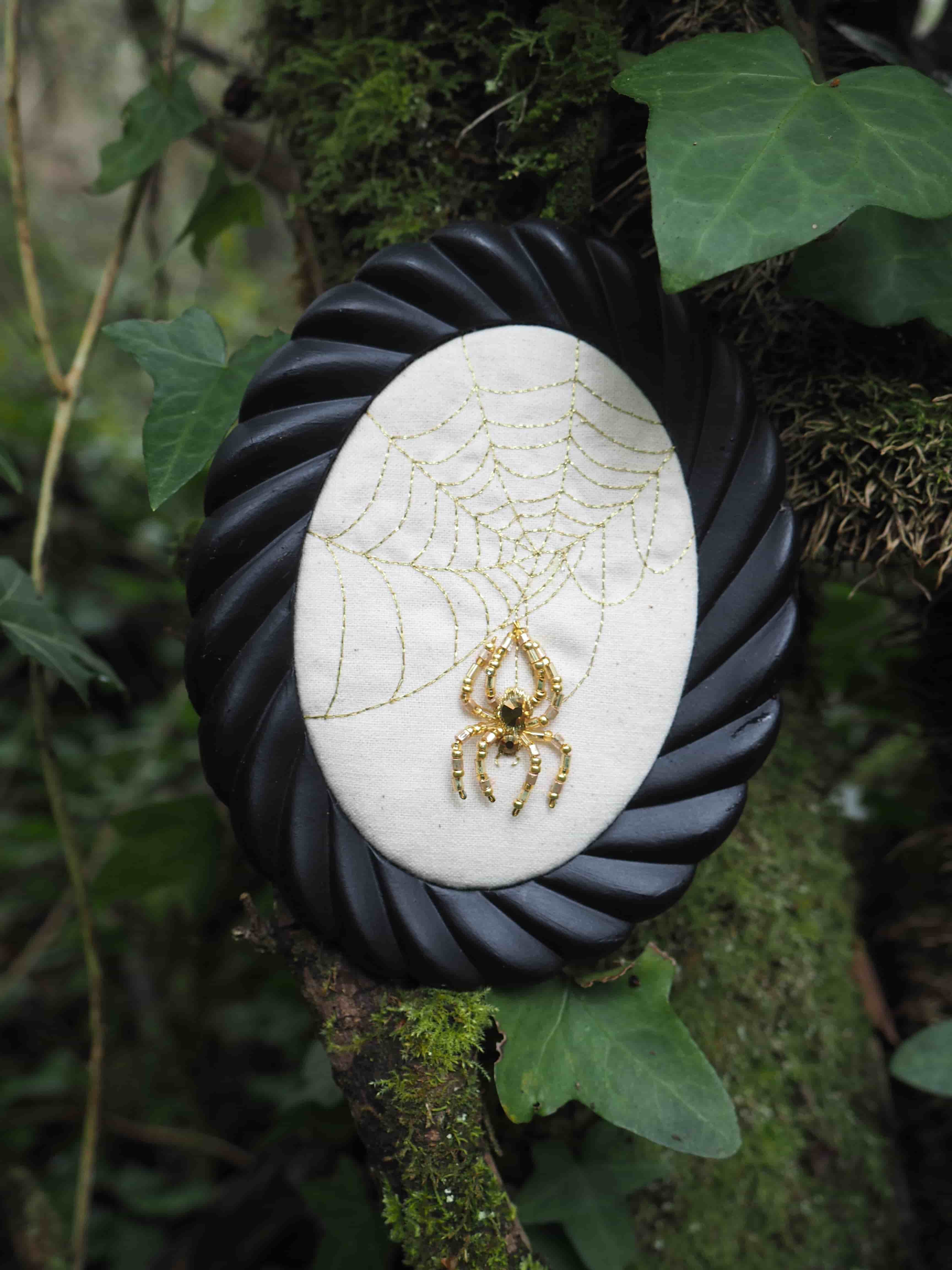 L'image présente un cadre ovale noir avec un motif torsadé, suspendu ou posé sur un tronc d'arbre couvert de mousse et de lierre. À l'intérieur du cadre, sur un fond de tissu blanc cassé, est brodée une toile d'araignée dorée. Au centre de cette toile, une broche en forme d'araignée dorée, ornée de ce qui semble être une petite pierre noire ou un cabochon au niveau de son corps, est épinglée. L'arrière-plan est flou, mais on distingue clairement la texture du tronc d'arbre, la mousse verte et les feuilles de lierre qui encadrent le sujet principal. L'éclairage semble naturel, mettant en valeur les détails dorés de l'araignée et de la toile. L'ambiance générale est un mélange de nature et d'un élément décoratif, avec une touche légèrement mystérieuse ou fantaisiste due au thème de l'araignée.