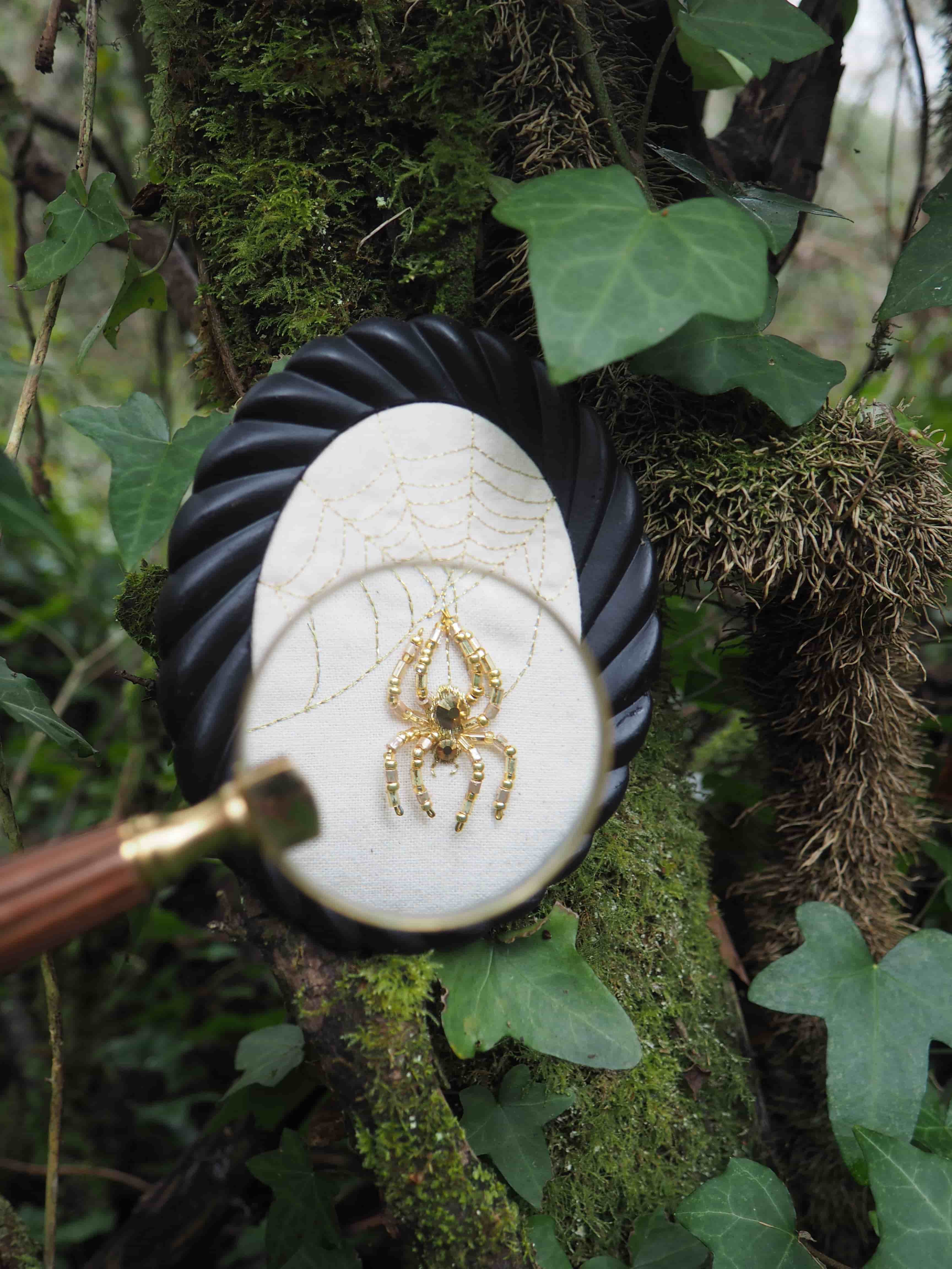 Cette image présente une scène intrigante et détaillée, centrée sur une broderie d'araignée observée à travers une loupe, le tout placé dans un environnement naturel. Au premier plan, une loupe est tenue, son manche en bois et en laiton s'étendant vers le bas à gauche. La lentille de la loupe est positionnée de manière à agrandir une partie de la broderie. Au centre de l'image, derrière la loupe, se trouve un petit cadre ovale noir, avec un motif en spirale ou torsadé sur son pourtour. À l'intérieur de ce cadre, une pièce de tissu blanc est visible. Sur ce tissu, une broderie délicate représente une toile d'araignée, avec des fils dorés ou de couleur claire formant les rayons et les spirales de la toile. Au centre de cette toile, une araignée est brodée avec des perles dorées et des pierres vertes, lui donnant un aspect précieux et détaillé. L'araignée est représentée avec ses huit pattes et son corps segmenté. L'arrière-plan est composé d'un tronc d'arbre ou d'une branche épaisse, recouverte de mousse verte et de lierre. Les feuilles de lierre, d'un vert profond, sont visibles à plusieurs endroits, ajoutant à l'ambiance naturelle et un peu mystérieuse de la scène. La texture de l'écorce de l'arbre est rugueuse et irrégulière, contrastant avec la finesse de la broderie. La lumière semble naturelle et diffuse, éclairant la scène de manière douce. L'ensemble de l'image suggère une exploration minutieuse de la nature et de l'artisanat, avec un focus particulier sur les détails.
