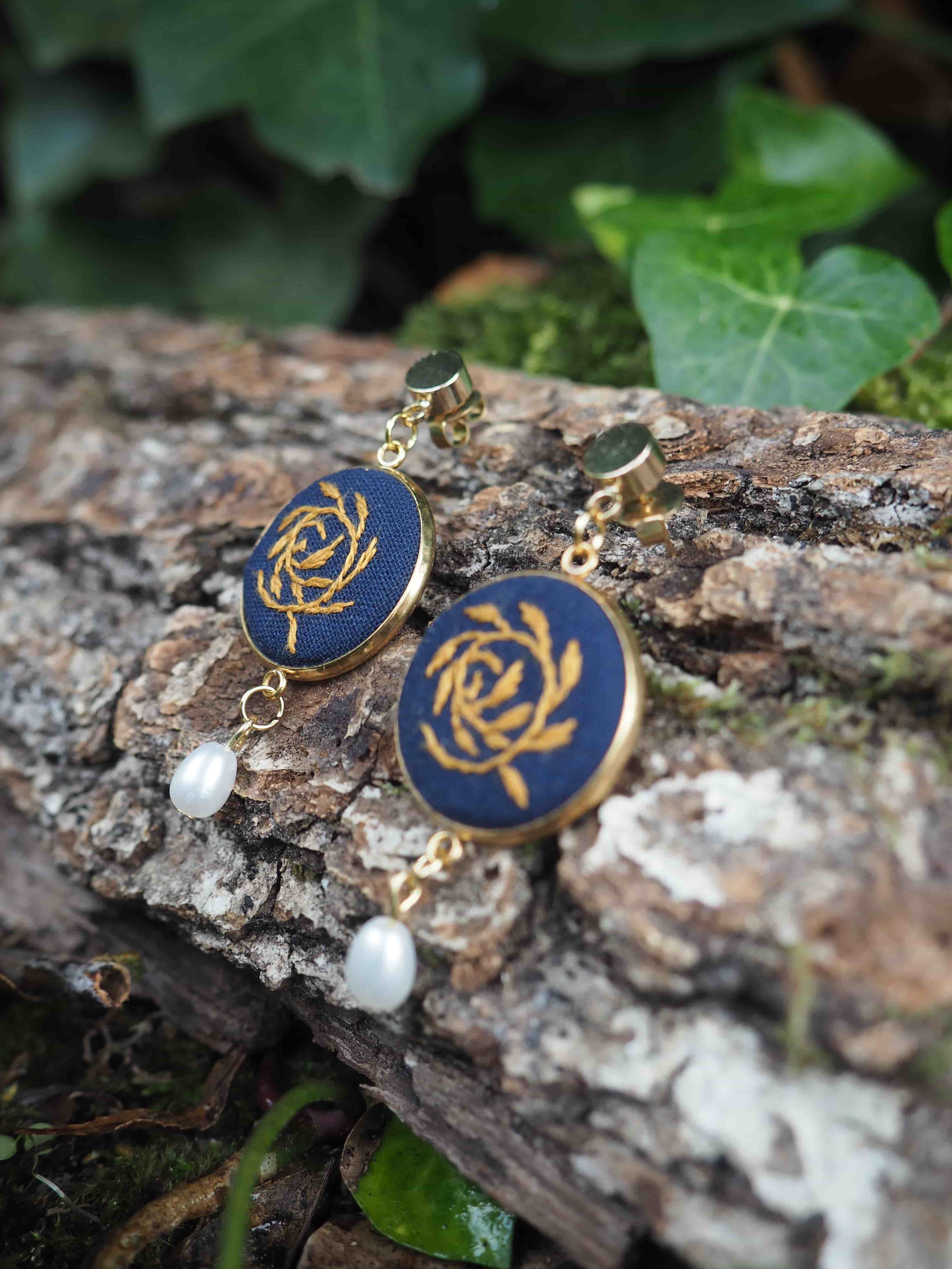 Cette image présente une paire de boucles d'oreilles élégantes posées sur un morceau de bois naturel dans un environnement extérieur verdoyant. **Description des bijoux :**
- Boucles d'oreilles pendantes avec des crochets dorés
- Médaillons circulaires de couleur bleu marine/bleu foncé
- Motif décoratif doré au centre de chaque médaillon, ressemblant à une rosace ou un motif floral stylisé
- Perles blanches nacrées suspendues en pendentif sous chaque médaillon **Cadre et composition :**
- Les bijoux reposent sur une surface de bois brut, avec une texture naturelle visible
- Arrière-plan flou montrant de la végétation verte (feuilles)
- Éclairage naturel qui met en valeur les reflets dorés et la brillance des perles
- Composition artistique qui crée un contraste harmonieux entre les éléments naturels et les bijoux raffinés L'ensemble dégage une esthétique à la fois naturelle et sophistiquée, mettant en valeur le travail artisanal des boucles d'oreilles dans un cadre organique.