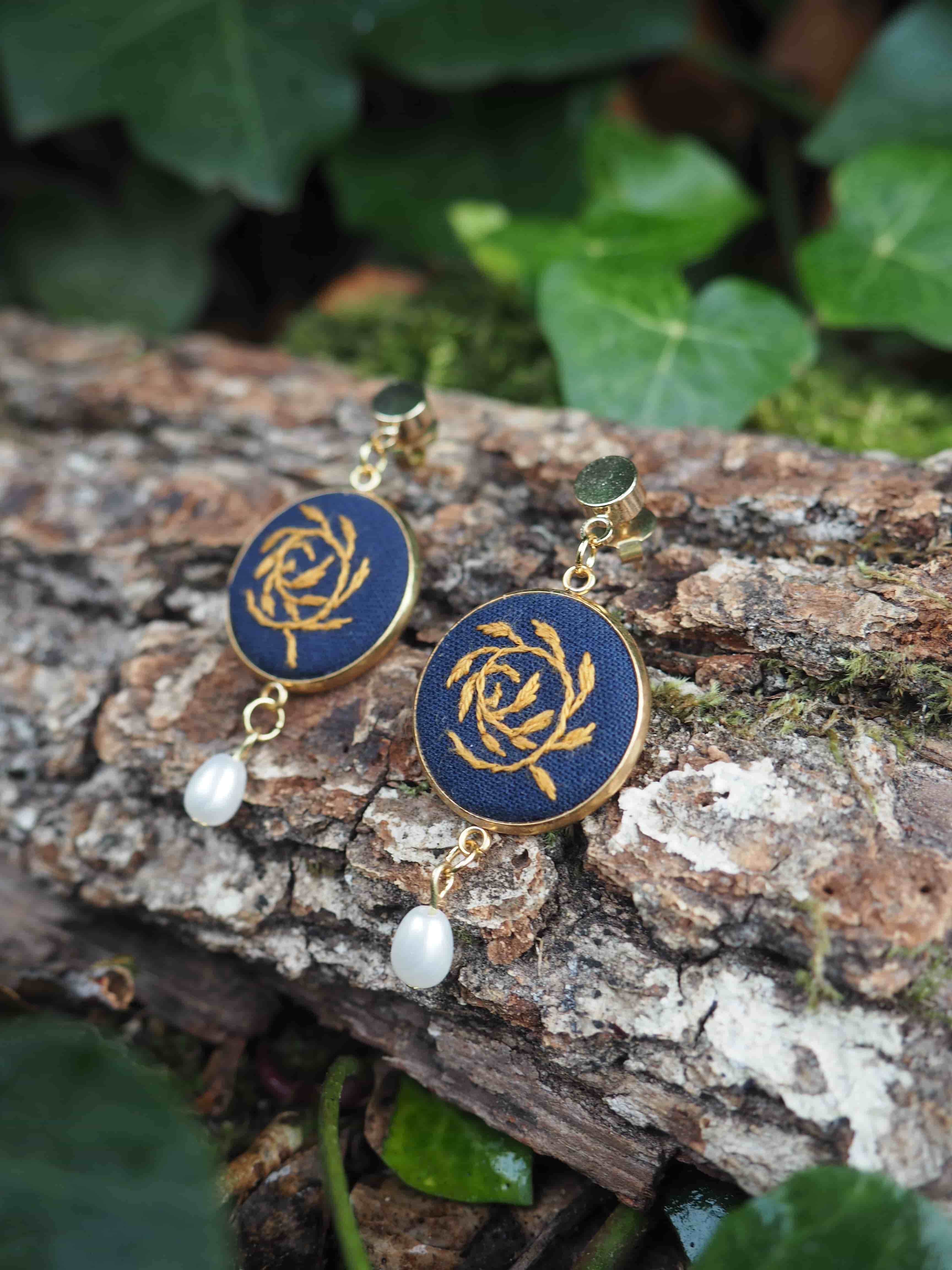 Cette image présente une paire de boucles d'oreilles élégantes posées sur un tronc d'arbre dans un environnement naturel.  **Caractéristiques des bijoux :**
- Boucles d'oreilles pendantes avec des crochets dorés
- Médaillons circulaires bleu marine ornés d'un motif doré en spirale ou rosace
- Petites perles blanches suspendues en pendentif sous chaque médaillon
- Style raffiné mêlant couleurs classiques (bleu, or, blanc) **Cadre naturel :**
- Les bijoux reposent sur l'écorce rugueuse d'un tronc d'arbre
- Présence de mousse ou lichen blanchâtre sur le bois
- Arrière-plan composé de feuillage vert luxuriant légèrement flou
- Éclairage naturel doux qui met en valeur les détails des bijoux Cette mise en scène crée un contraste harmonieux entre l'élégance des bijoux et la rusticité de l'environnement forestier, donnant une dimension organique et authentique à la présentation de ces accessoires de mode.