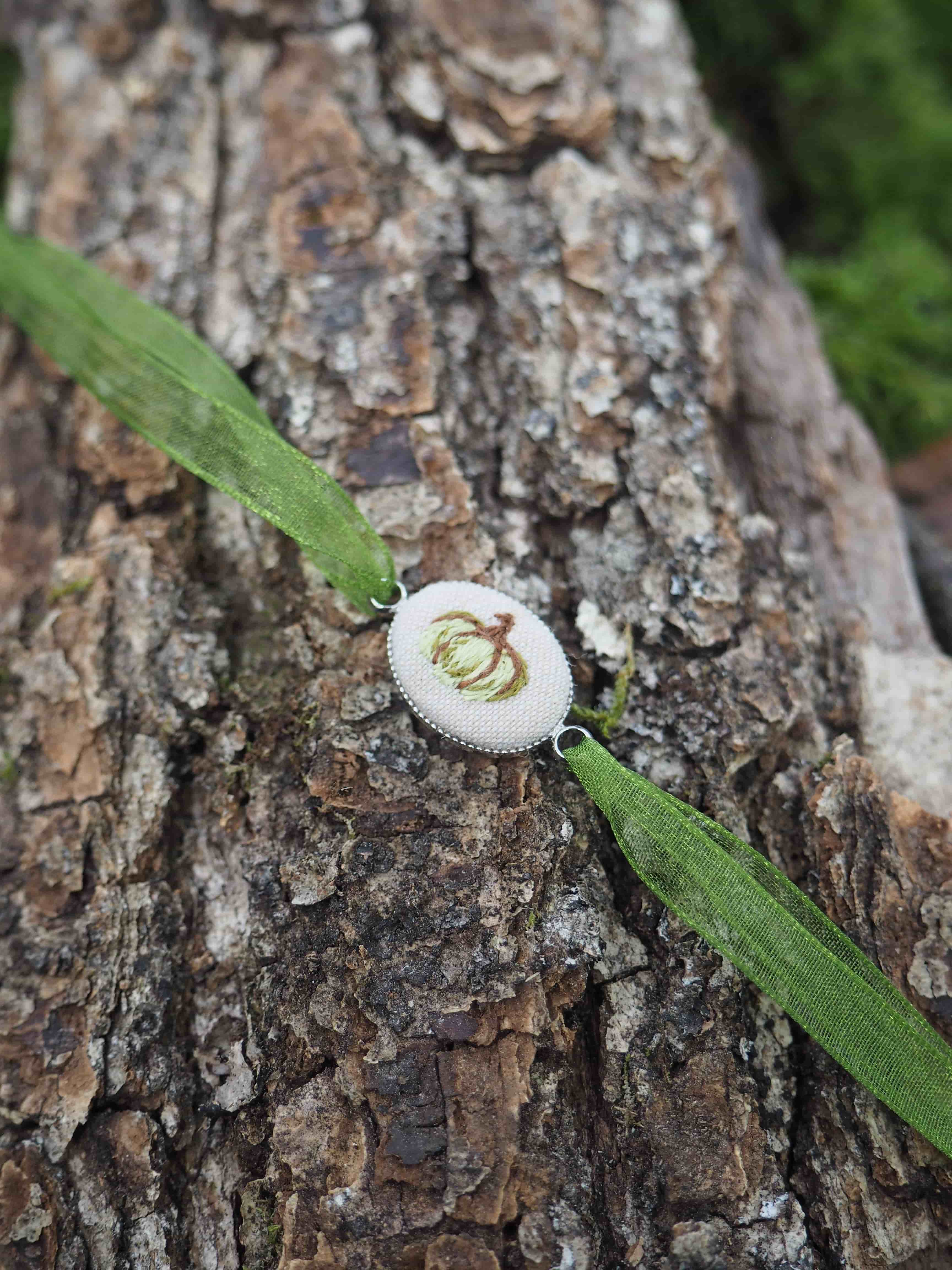 Cette image présente une composition naturelle délicate sur l'écorce rugueuse d'un arbre. Au centre de la photo se trouve un petit champignon blanc de forme circulaire, probablement un polypore ou un champignon bracket, avec une surface lisse et claire qui contraste avec la texture brune et crevassée de l'écorce. Le champignon est traversé par ce qui semble être un brin d'herbe ou une feuille étroite de couleur vert vif, créant une ligne diagonale qui divise harmonieusement la composition. Cette herbe ou cette feuille ajoute une touche de couleur fraîche à la palette autrement neutre de bruns, de gris et de blanc. L'écorce de l'arbre présente une texture riche et détaillée, avec des fissures, des crevasses et des variations de couleur allant du brun foncé au gris clair. En arrière-plan, on distingue un flou artistique de verdure qui suggère un environnement forestier. L'éclairage naturel met en valeur les textures et crée une atmosphère paisible et contemplative, typique d'une observation attentive de la nature dans son habitat naturel.