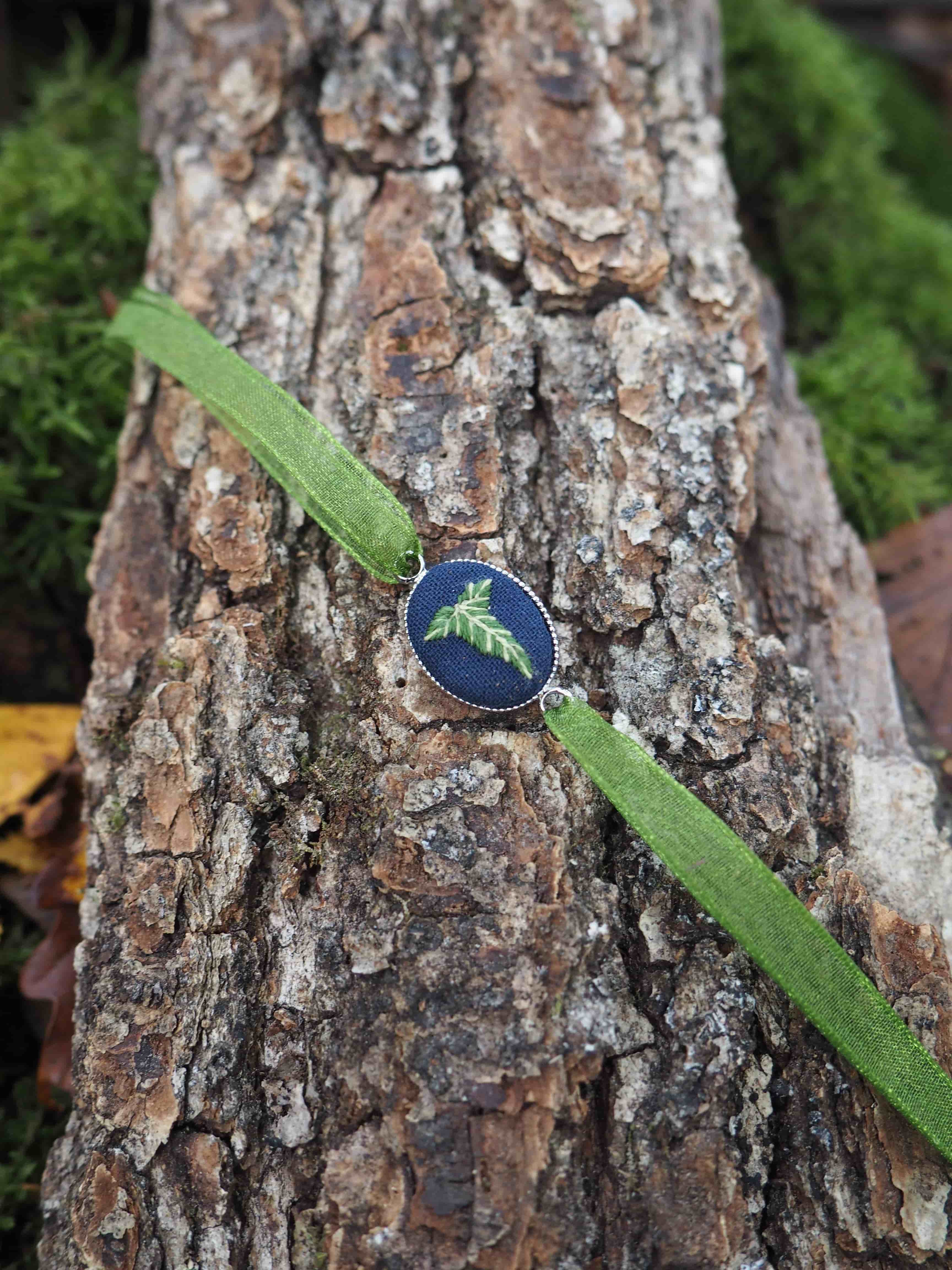 Cette image présente une composition naturelle très esthétique. Au centre, on observe un petit pendentif ou bijou de forme ovale avec une surface bleu-vert iridescente qui rappelle les couleurs d'une aile de papillon ou d'un scarabée. Ce bijou est délicatement posé sur un ruban ou une bande de tissu vert olive qui traverse diagonalement l'image. L'ensemble repose sur un tronc d'arbre ou une branche dont l'écorce rugueuse et texturée présente des tons bruns et gris, avec des motifs naturels créés par les fissures et les reliefs du bois. En arrière-plan, on distingue un tapis de mousse d'un vert vif qui apporte une belle profondeur de champ et fait ressortir les éléments du premier plan. La photographie capture bien les contrastes entre les différentes textures - la douceur du tissu, la rugosité de l'écorce et le velouté de la mousse - ainsi que la palette de couleurs naturelles dominée par les verts et les bruns, avec cette touche d'éclat iridescent du bijou qui attire immédiatement le regard.