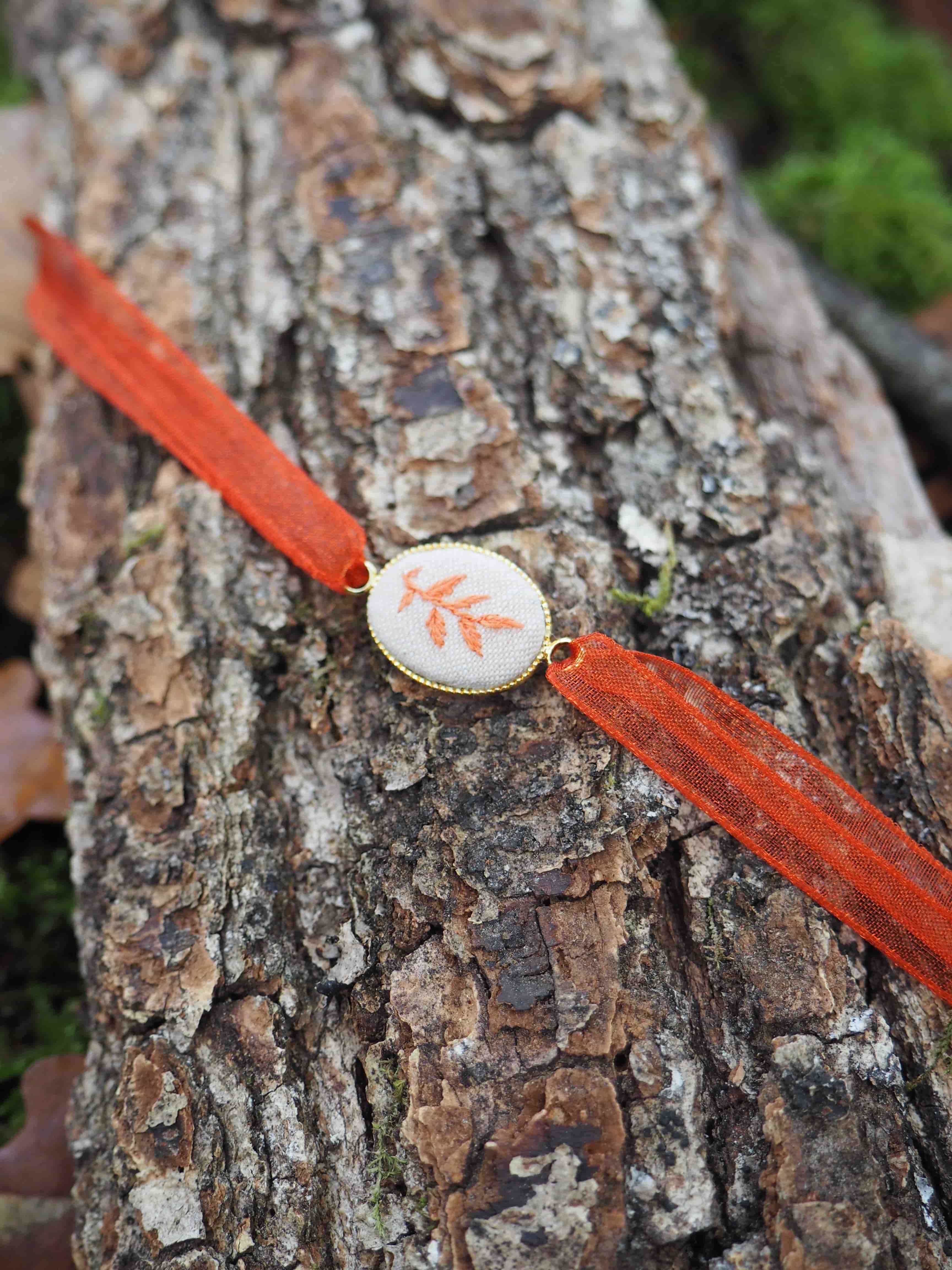 Cette image montre un bracelet ou un accessoire décoratif posé sur l'écorce rugueuse d'un tronc d'arbre. Le bracelet présente les caractéristiques suivantes : **Éléments principaux :**
- Un ruban ou cordon de couleur orange/rouge vif
- Un médaillon central rond de couleur blanche ou crème
- Sur le médaillon, on distingue un motif décoratif qui ressemble à une feuille ou un motif floral de couleur orange/corail **Contexte :**
- L'objet est photographié sur l'écorce naturelle d'un arbre, avec sa texture caractéristique rugueuse et ses tons bruns-gris
- L'arrière-plan est légèrement flou, laissant apercevoir de la végétation verte
- L'éclairage naturel met en valeur les contrastes entre les couleurs vives du bracelet et les tons neutres de l'écorce **Style :**
L'ensemble évoque un accessoire artisanal ou fait main, avec un style naturel et bohème qui s'harmonise bien avec l'environnement forestier dans lequel il est photographié.