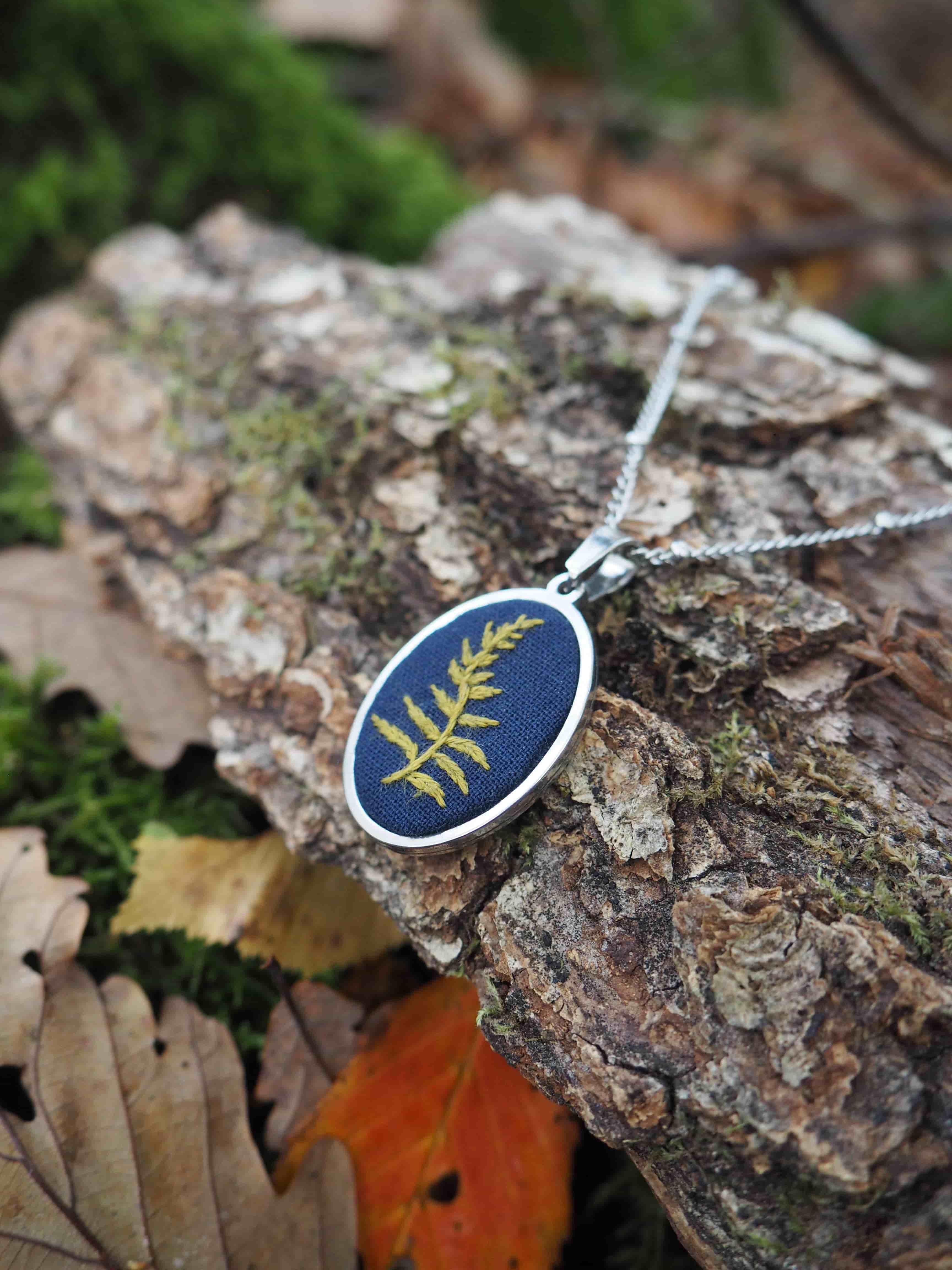 Cette image présente un bijou artisanal posé sur un tronc d'arbre dans un environnement naturel. Il s'agit d'un pendentif ovale avec un fond bleu marine sur lequel est délicatement incrustée une feuille de fougère dorée, créant un contraste élégant. Le pendentif est suspendu à une chaîne argentée fine. Le bijou repose sur l'écorce rugueuse et texturée d'un tronc d'arbre, entouré de feuilles d'automne aux teintes chaudes - des feuilles brunes, orangées et quelques feuilles vertes encore fraîches. L'arrière-plan est artistiquement flouté, laissant deviner une végétation luxuriante avec des tons verts. Cette composition photographique met en valeur le caractère naturel et organique du bijou, créant une harmonie parfaite entre l'objet artisanal et son environnement forestier. L'éclairage naturel fait ressortir les détails de la feuille dorée et accentue les textures de l'écorce et des feuilles environnantes.